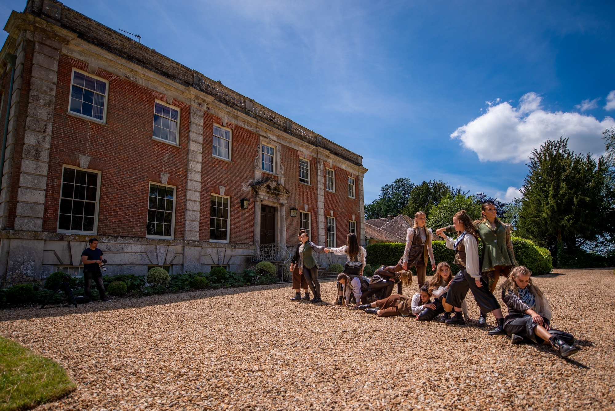 A group of Arts University Bournemouth student performers in period costume sitting on a gravel driveway outside a stately home-like building.