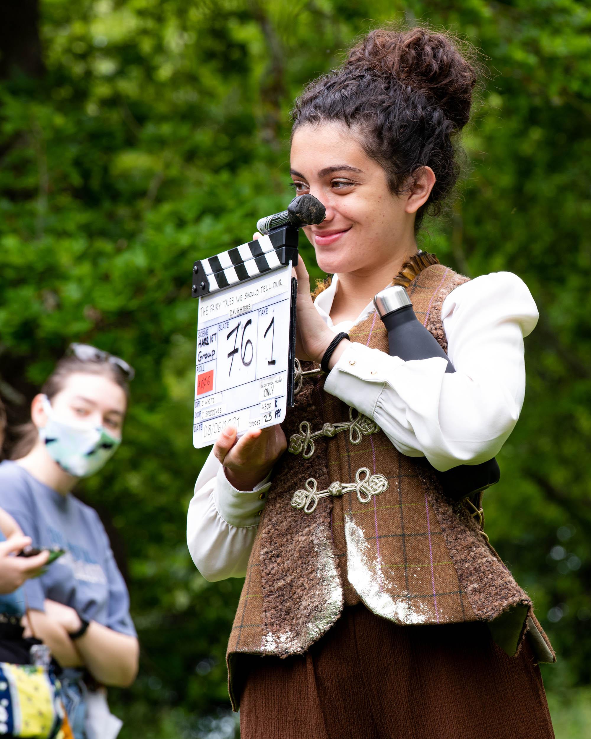 A person holding a clapper board. There are trees and another person wearing a face mask in the background.