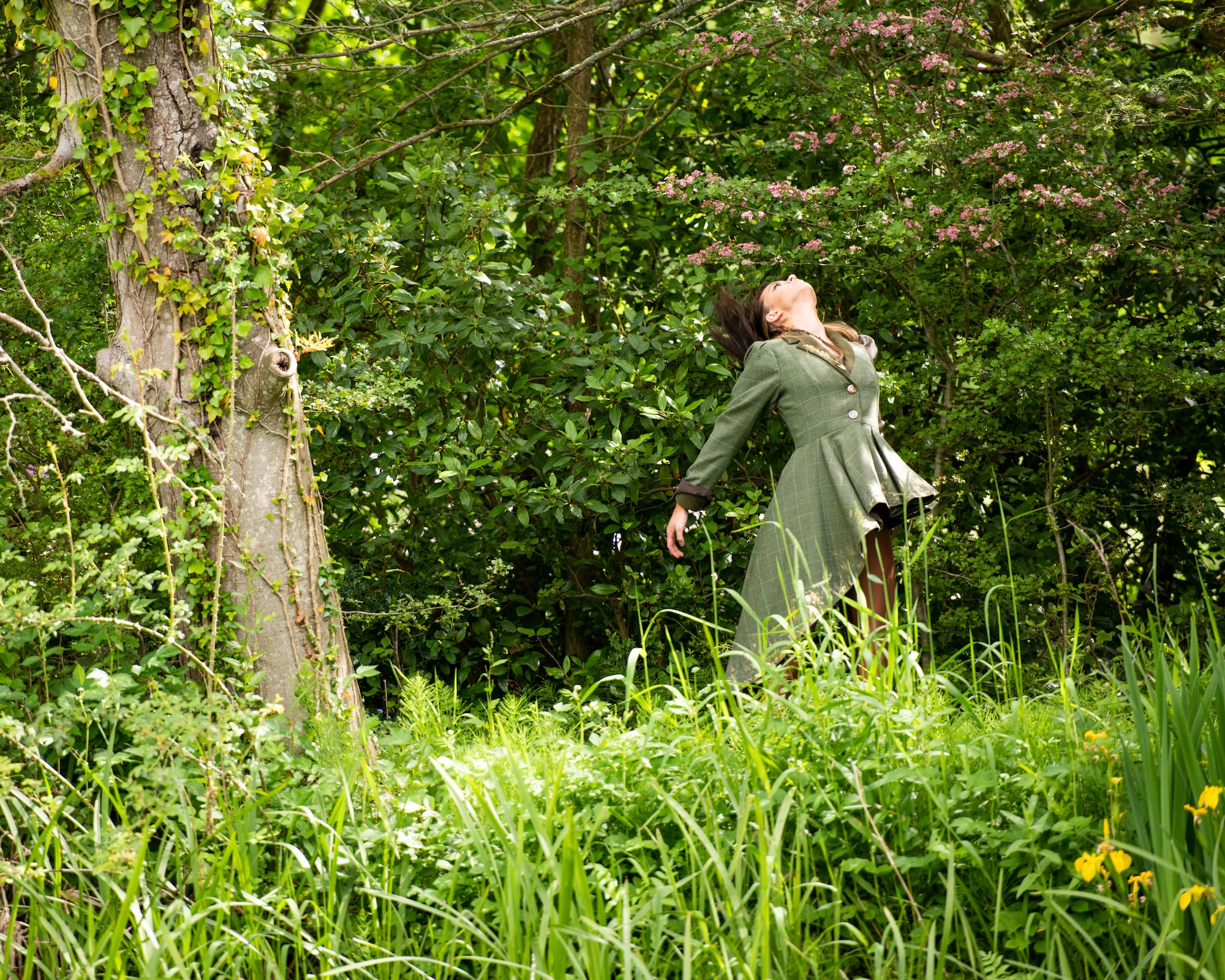 A woman in a green coat stands in tall grass with her head tilted back, surrounded by dense woodland and a tree trunk on the left.