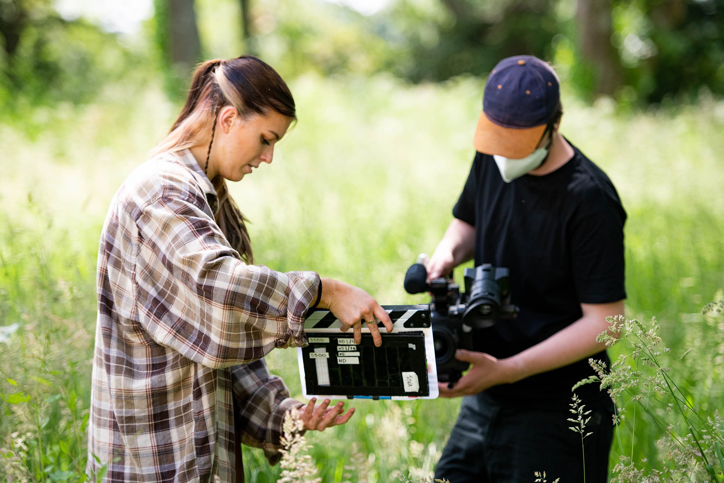 A person in a face mask looks down at a film camera; another person holds a film clapper board in front of it.