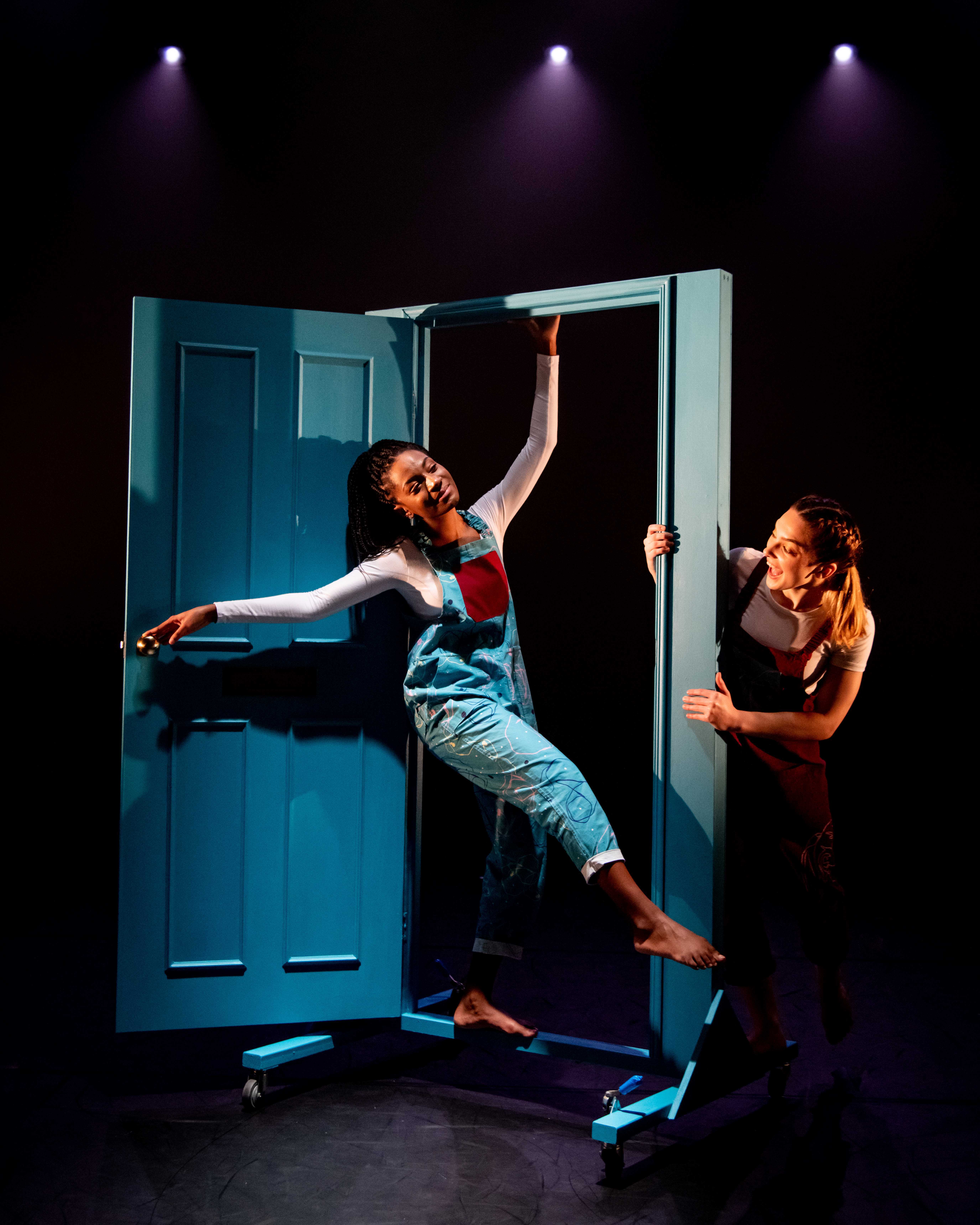 Two performers on stage with blue door frame prop under purple spotlights. One person hanging from door frame, another standing beside it.