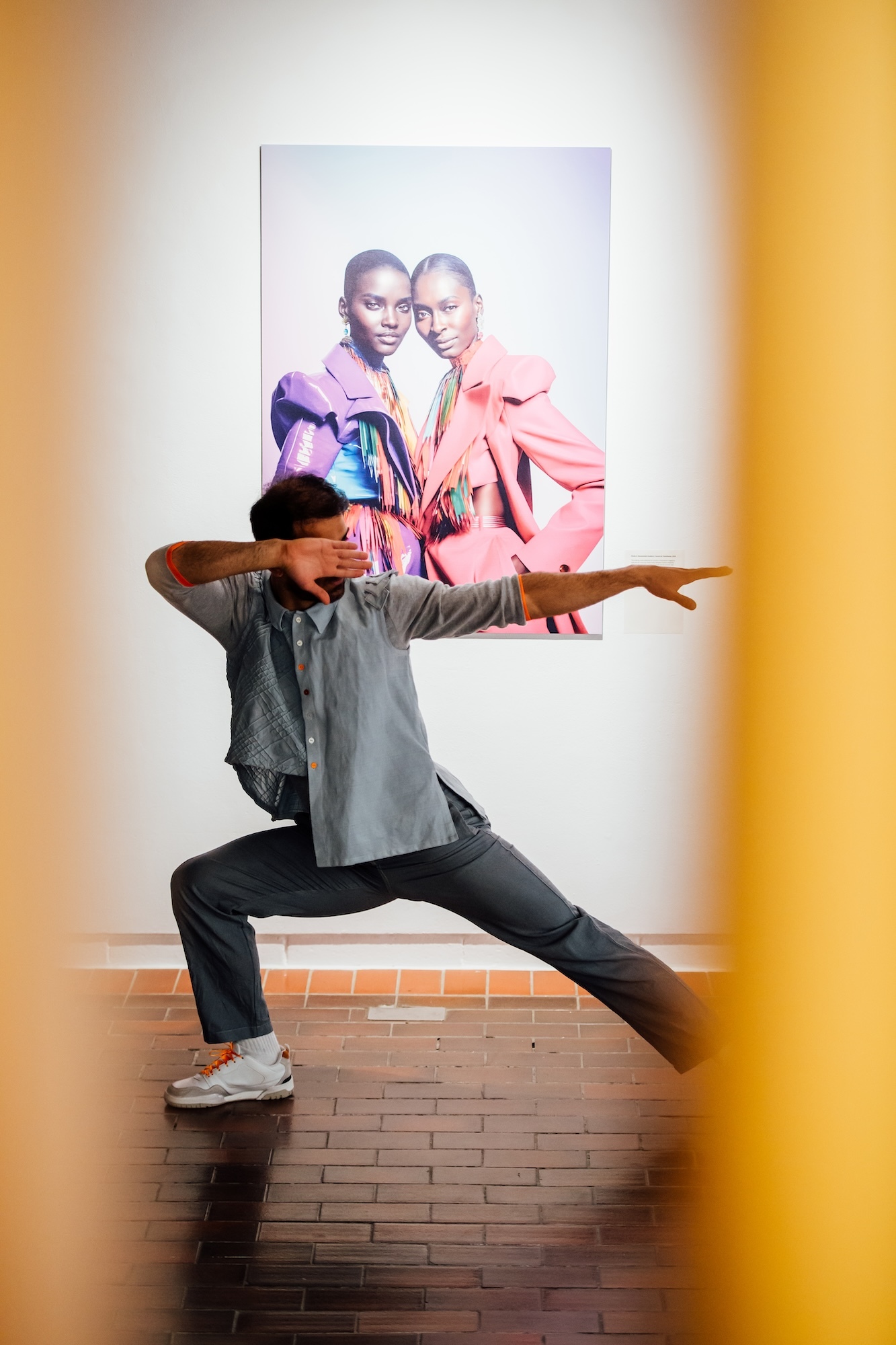A BA (Hons) Dance student posing with one arm and one leg stretched out, performing at TheGallery, Arts University Bournemouth. An AI-generated artwork of two people is on display behind them.