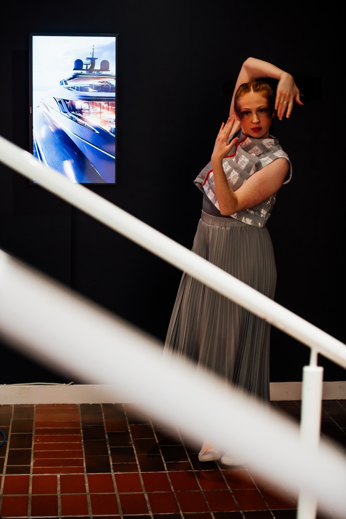 A BA (Hons) Dance student pulls a pose while dancing at TheGallery, Arts University Bournemouth. An AI-generated artwork of a yacht is on display to their left. The hand rail of a staircase is visible in the foreground.