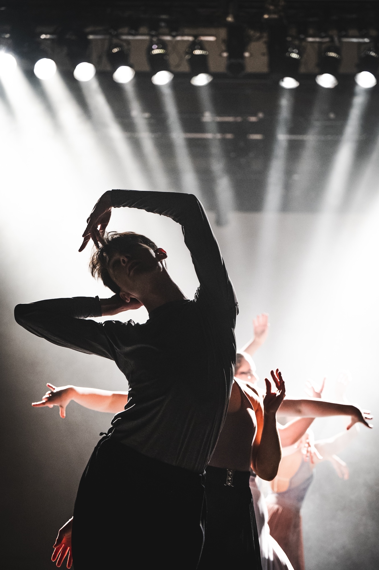 A BA (Hons) Dance student in partial silhouette, several spotlights shining down behind them. Their body is tilted towards the left and one arm raised over and touching their head.