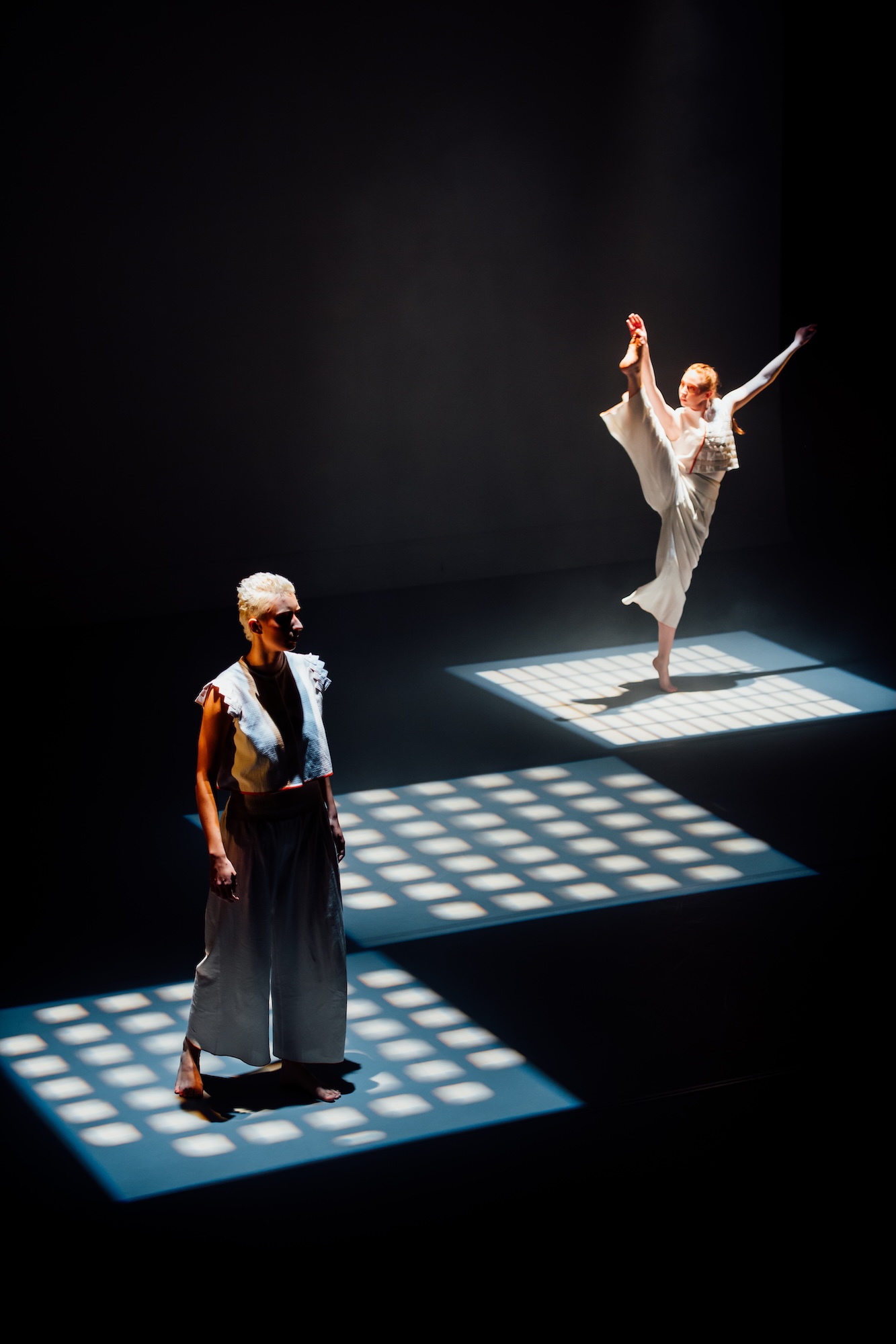 Two dancers on a dark stage, one standing in the foreground and the other balancing in a raised pose on a lit platform behind.