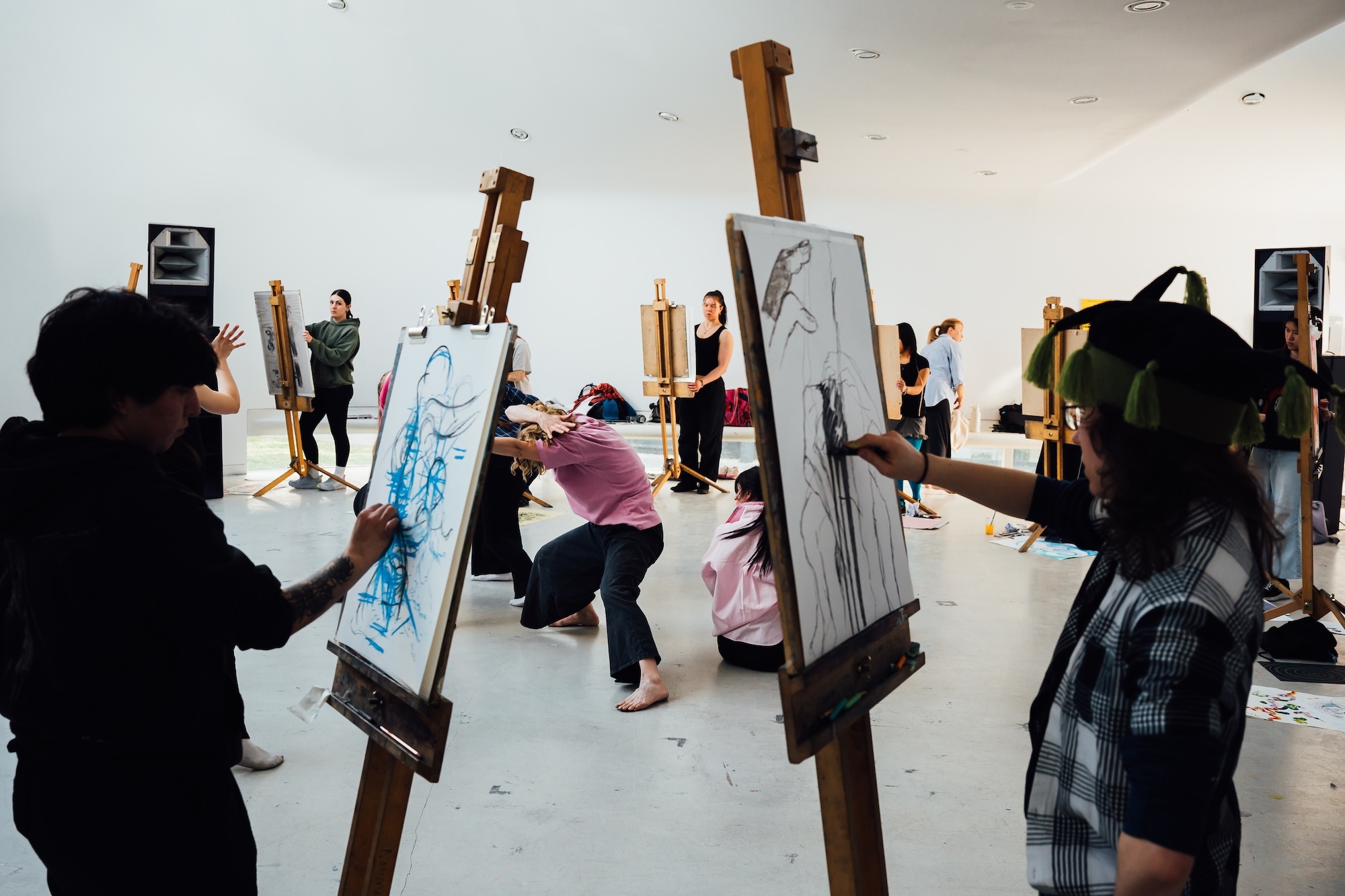 Art class with students painting at wooden easels in white room, instructor standing in background, various artworks on canvases
