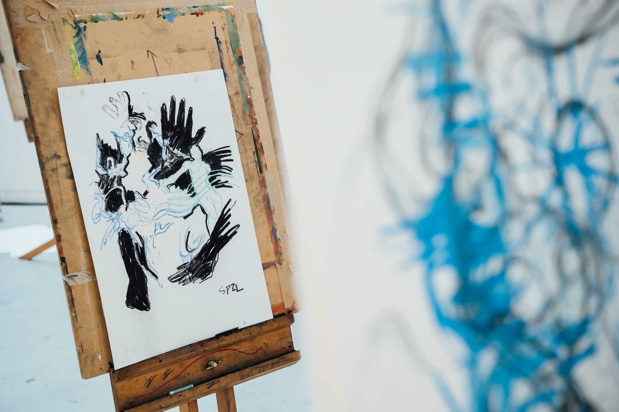 Black ink drawing of birds on white paper mounted on wooden easel, with blurred blue artwork visible in background