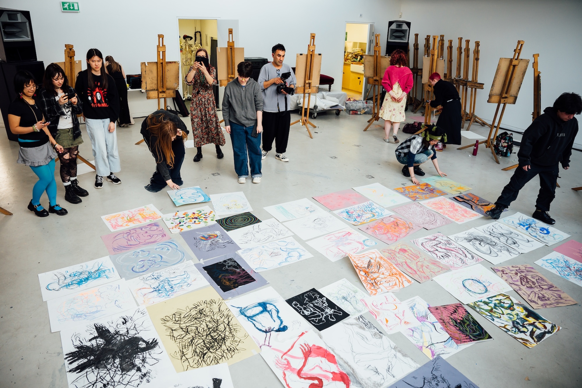 Students gathered around colourful artwork laid out on white floor in art studio with easels and equipment visible