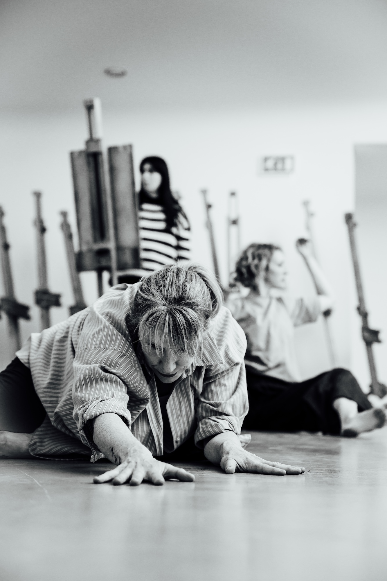 Woman kneeling on floor in dance studio, others stretching in background, ballet barres visible along walls, black and white image