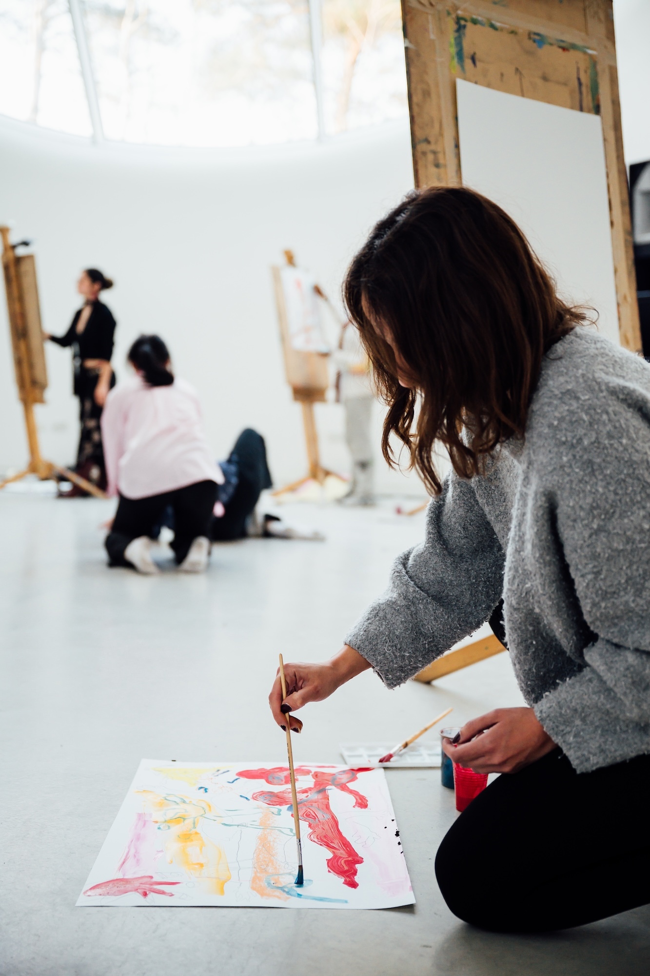 Woman in grey jumper kneeling on floor painting with brush on paper with pink and yellow colours in art studio