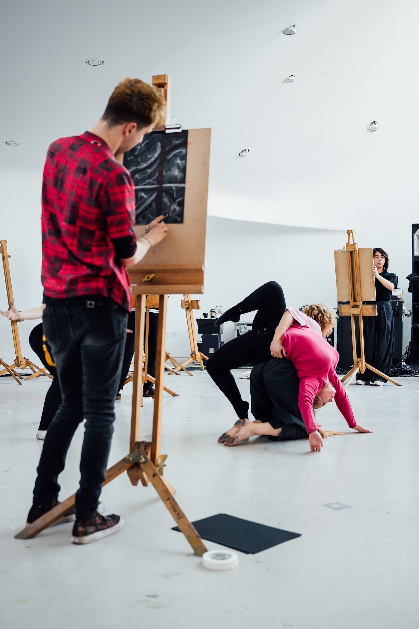 Man in red plaid shirt painting at easel whilst woman in pink top does yoga pose on floor in white art studio with wooden easels