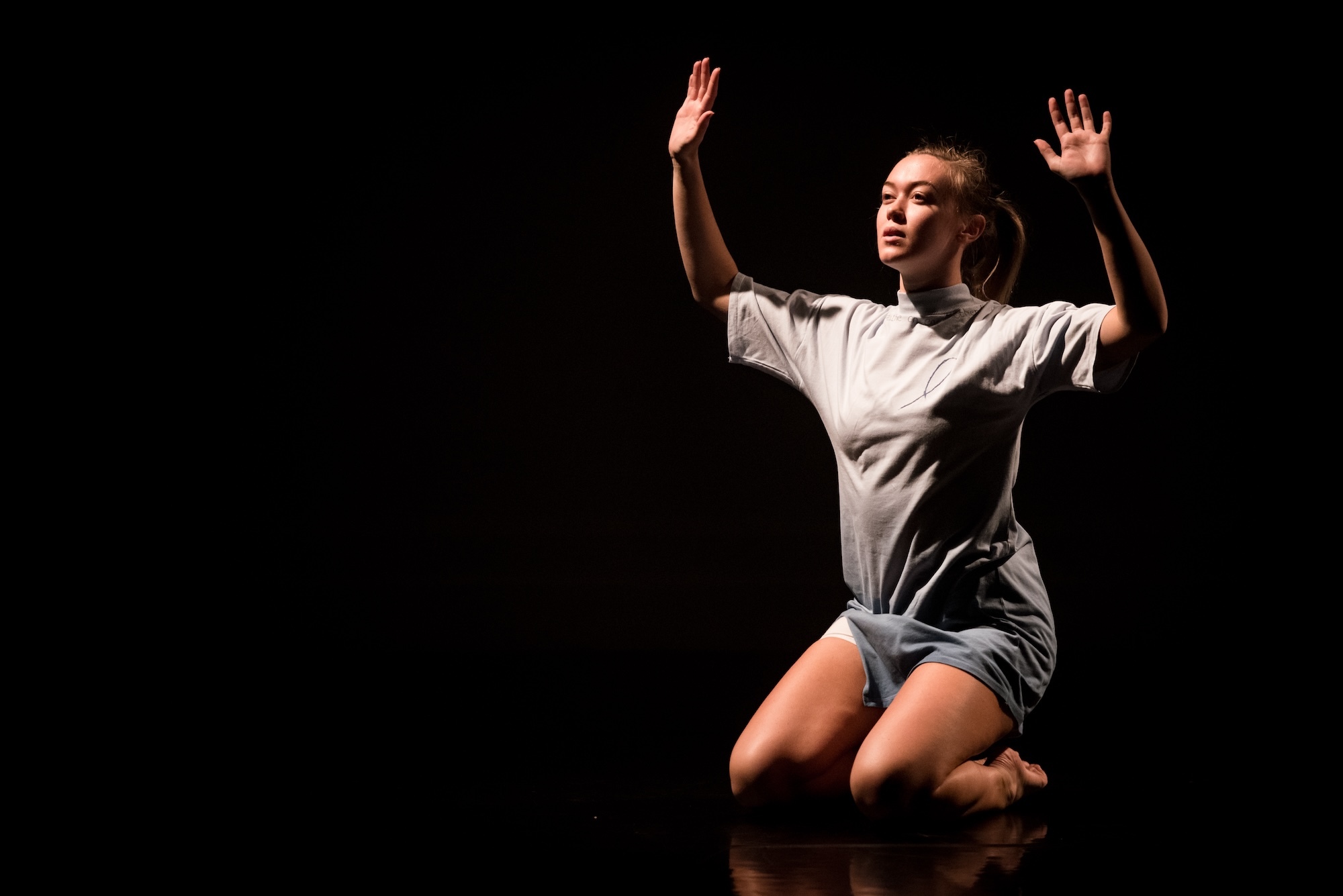 An Arts University Bournemouth student performer kneeling in a dark space with their arms and hands raised.