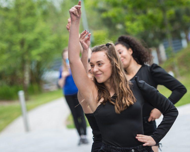 A line of dancers with their arms up, clicking their fingers, in the middle of a routine on a footpath.