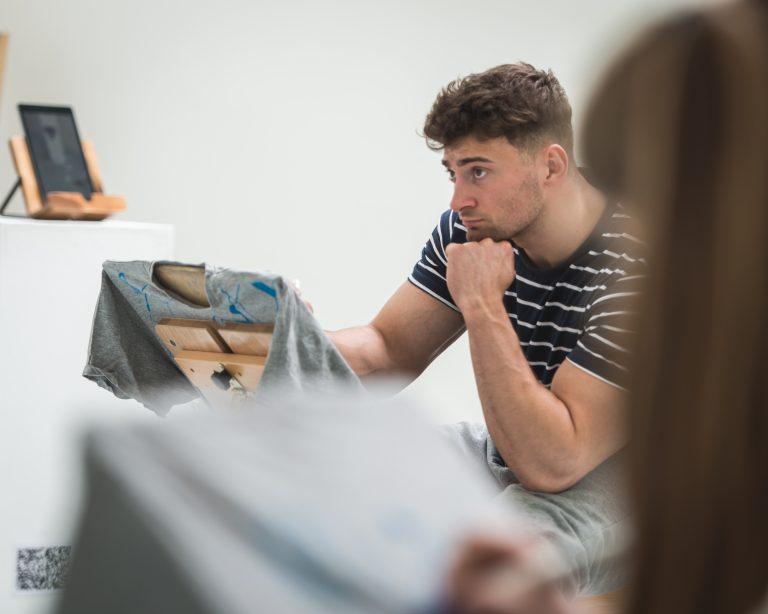 A person sits with their chin on their hand and paints on an easel.