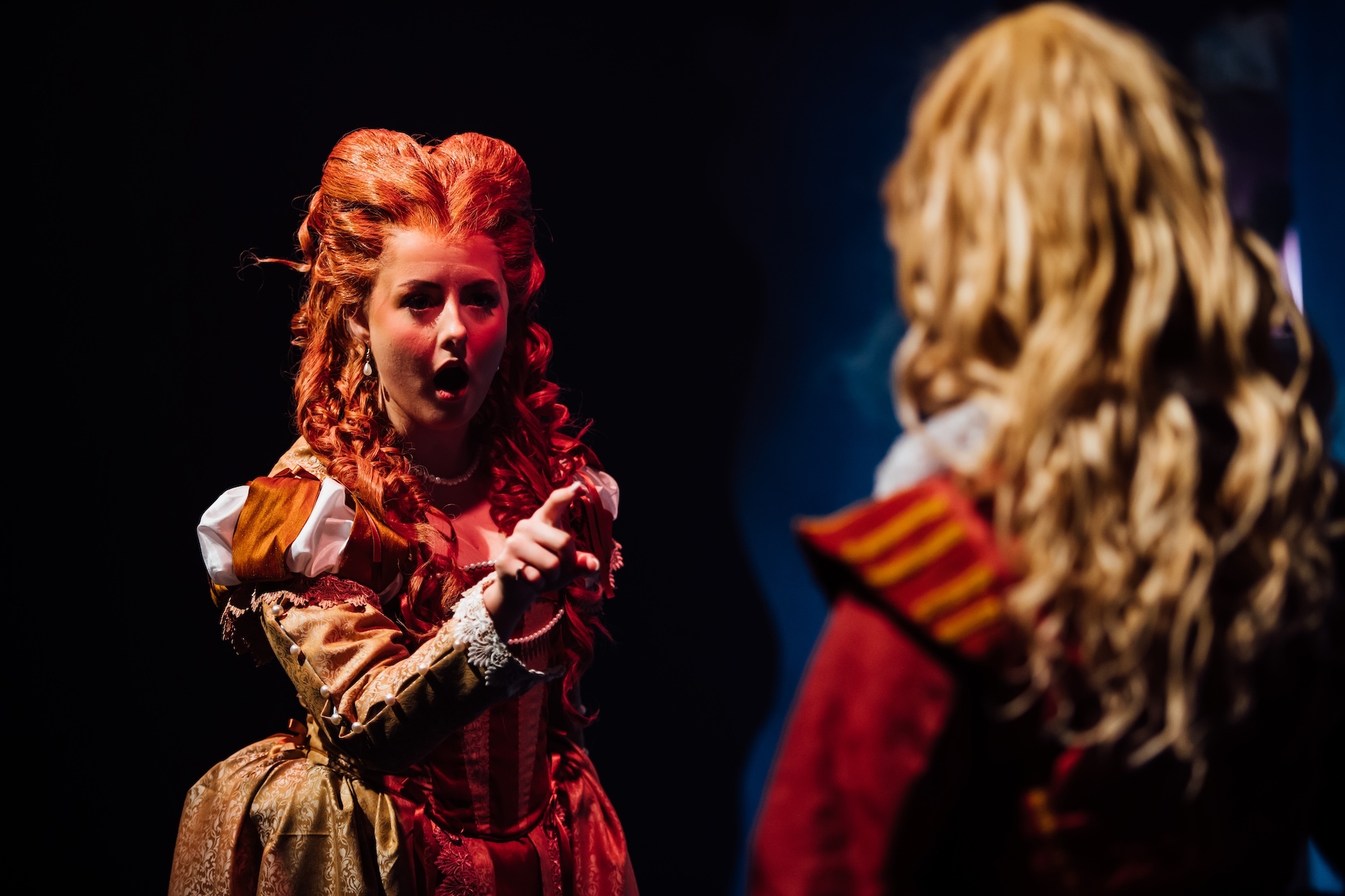 Two actors on stage in 17th Century dress wear, one pointing a finger at the other under red lighting.