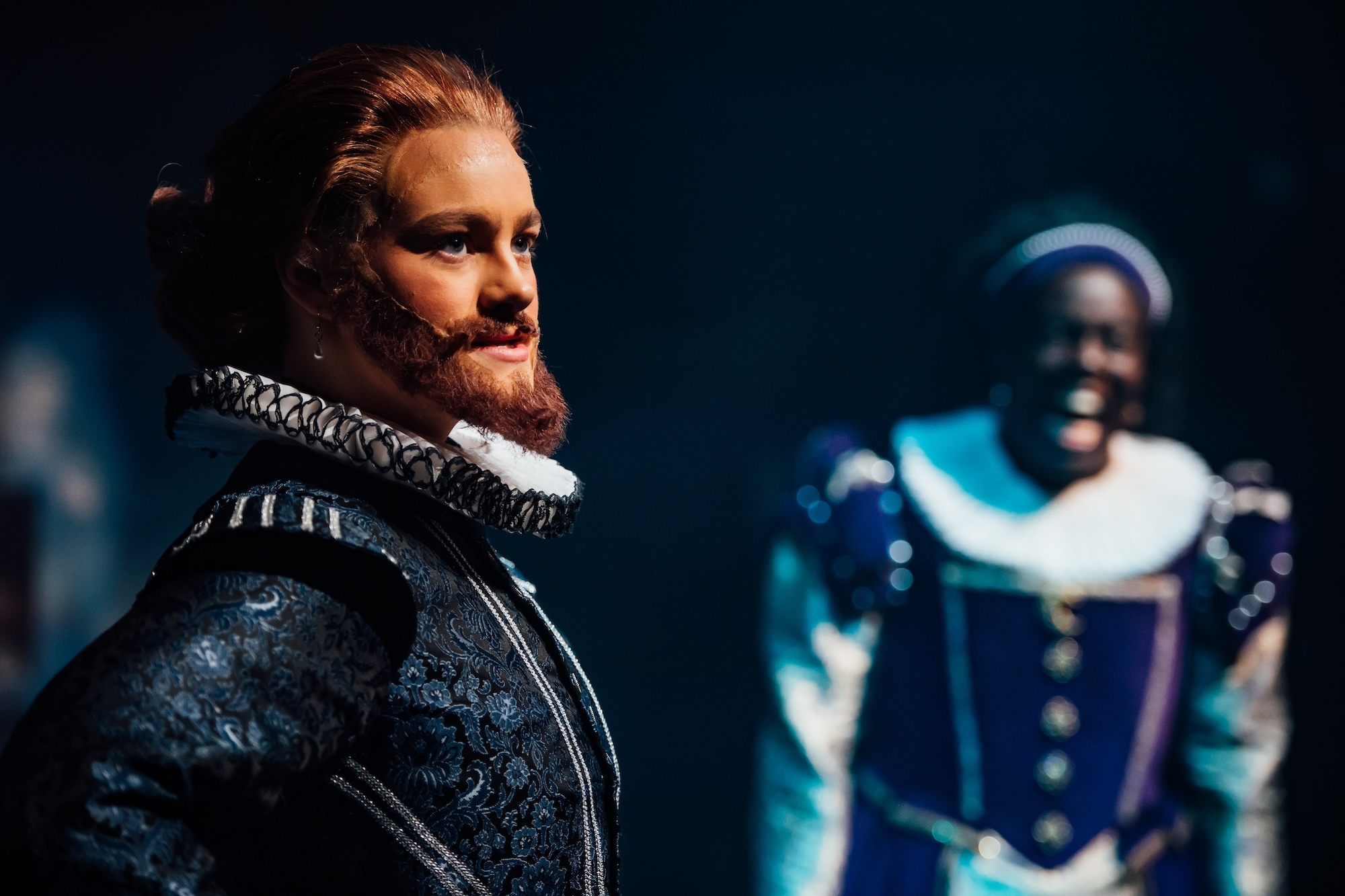 Two actors on stage in 17th Century garments, one under lighting, the other blurred in the background.