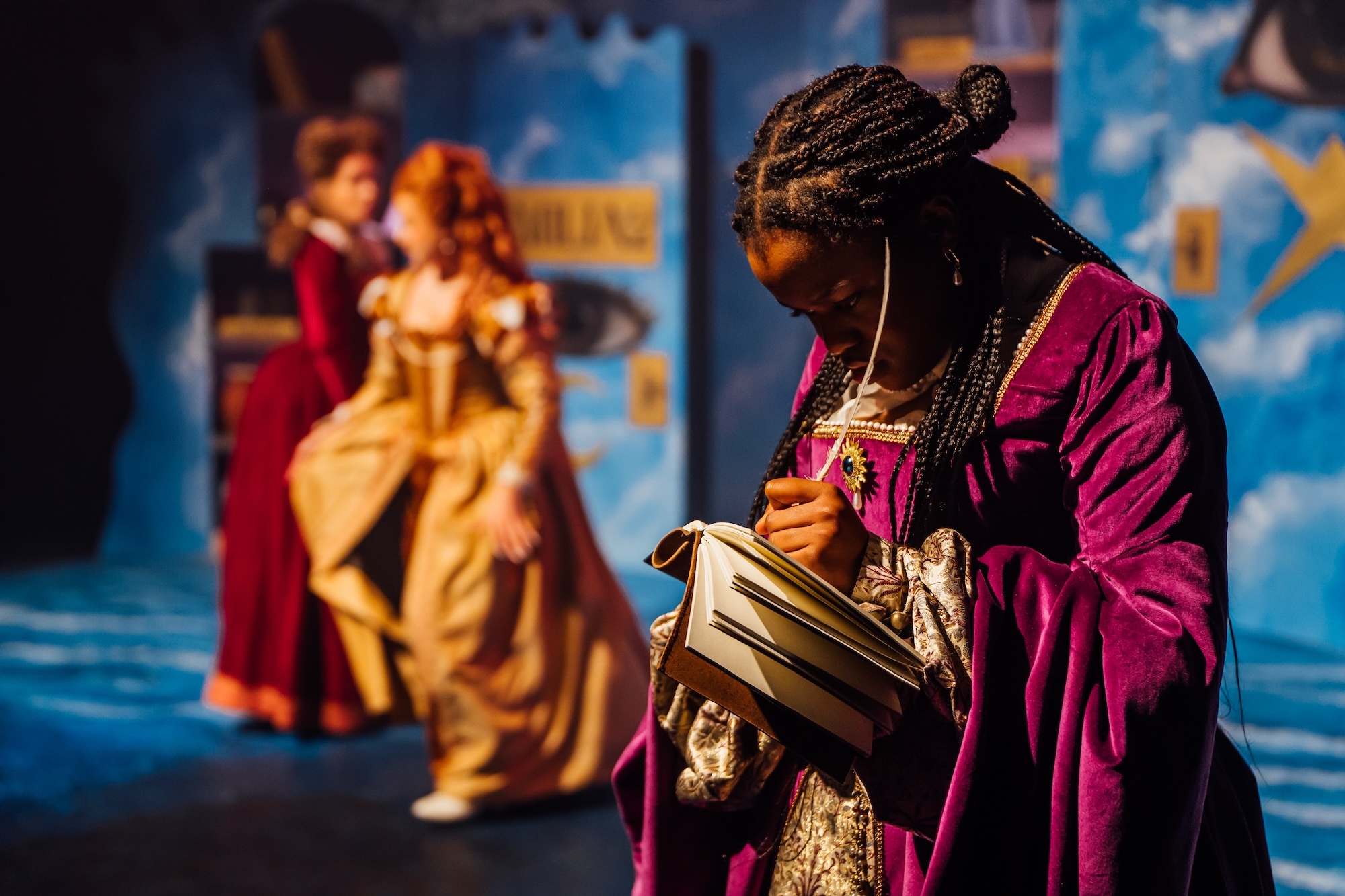 Actors in 17th century costume on stage, one in pink writing in a book, the others blurred in the background.