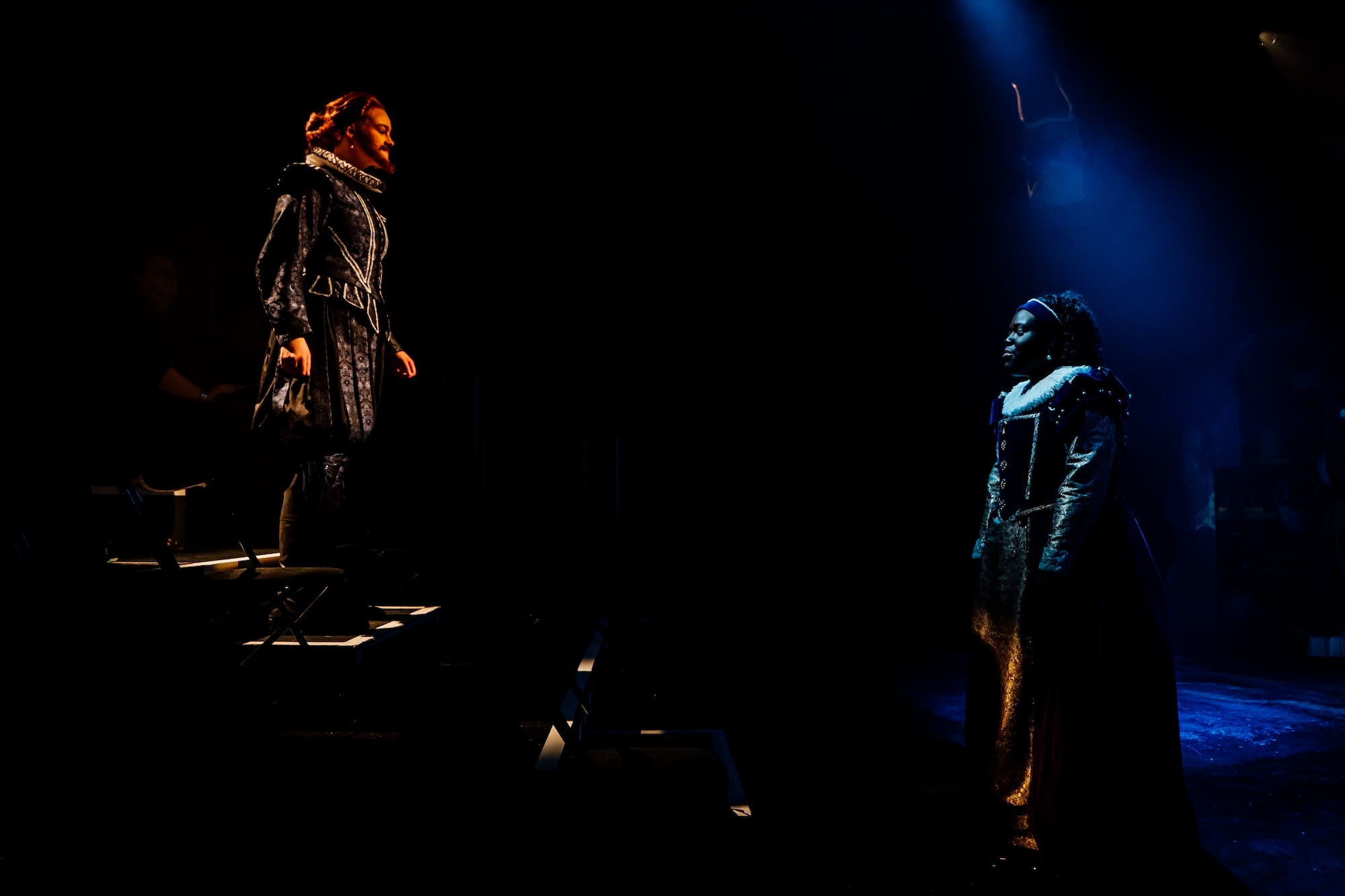 Two actors on stage in 17th Century costumes, one on stairs, the other under blue light