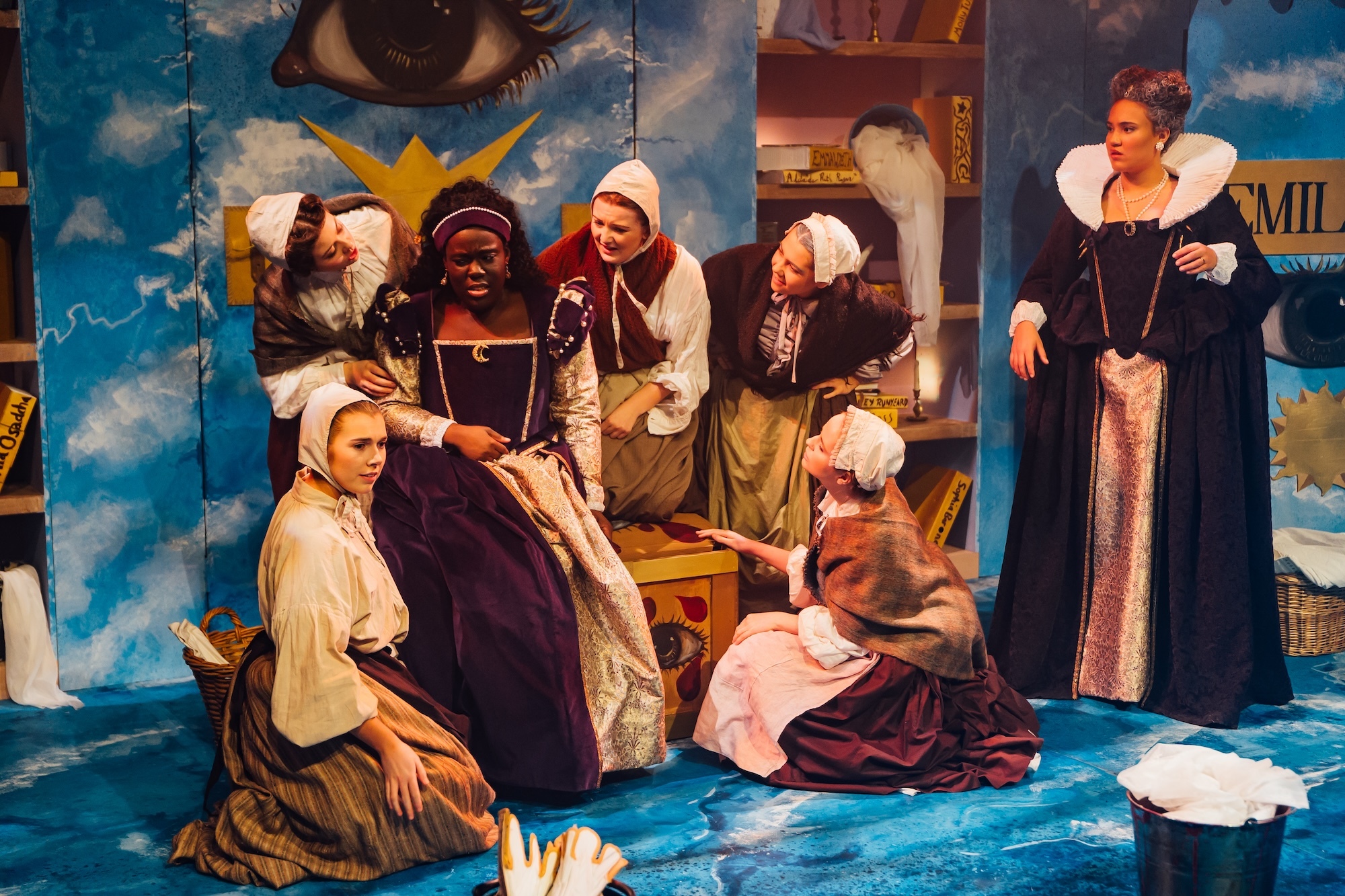 Actors on stage in 17th Century costume, abstract set design with clouds and eyes.