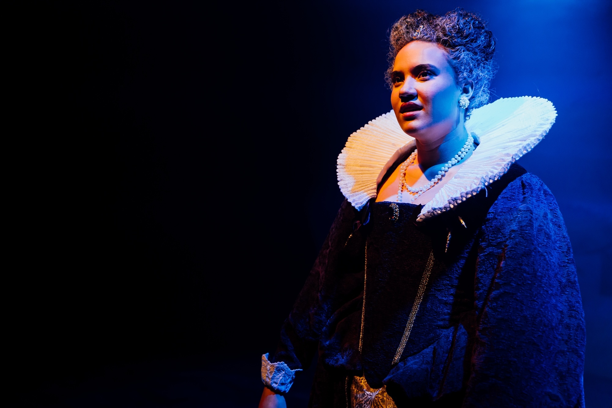 An actor under blue stage lighting in a 17th Century costume.