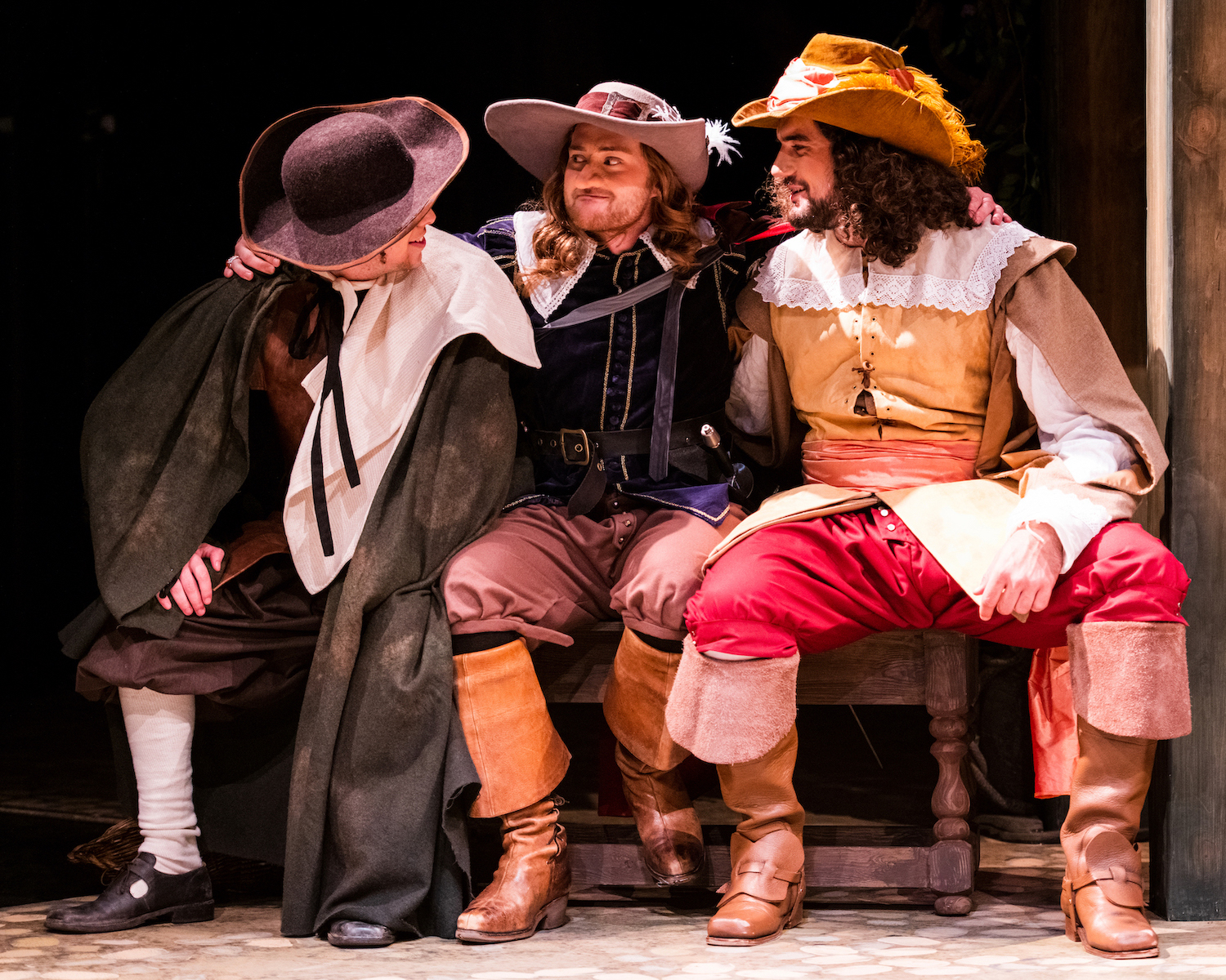 Three BA (Hons) Acting students in period costume sit next to each other on a bench, performing Cyrano de Bergerac. The actor in the centre has his arms around the other two.