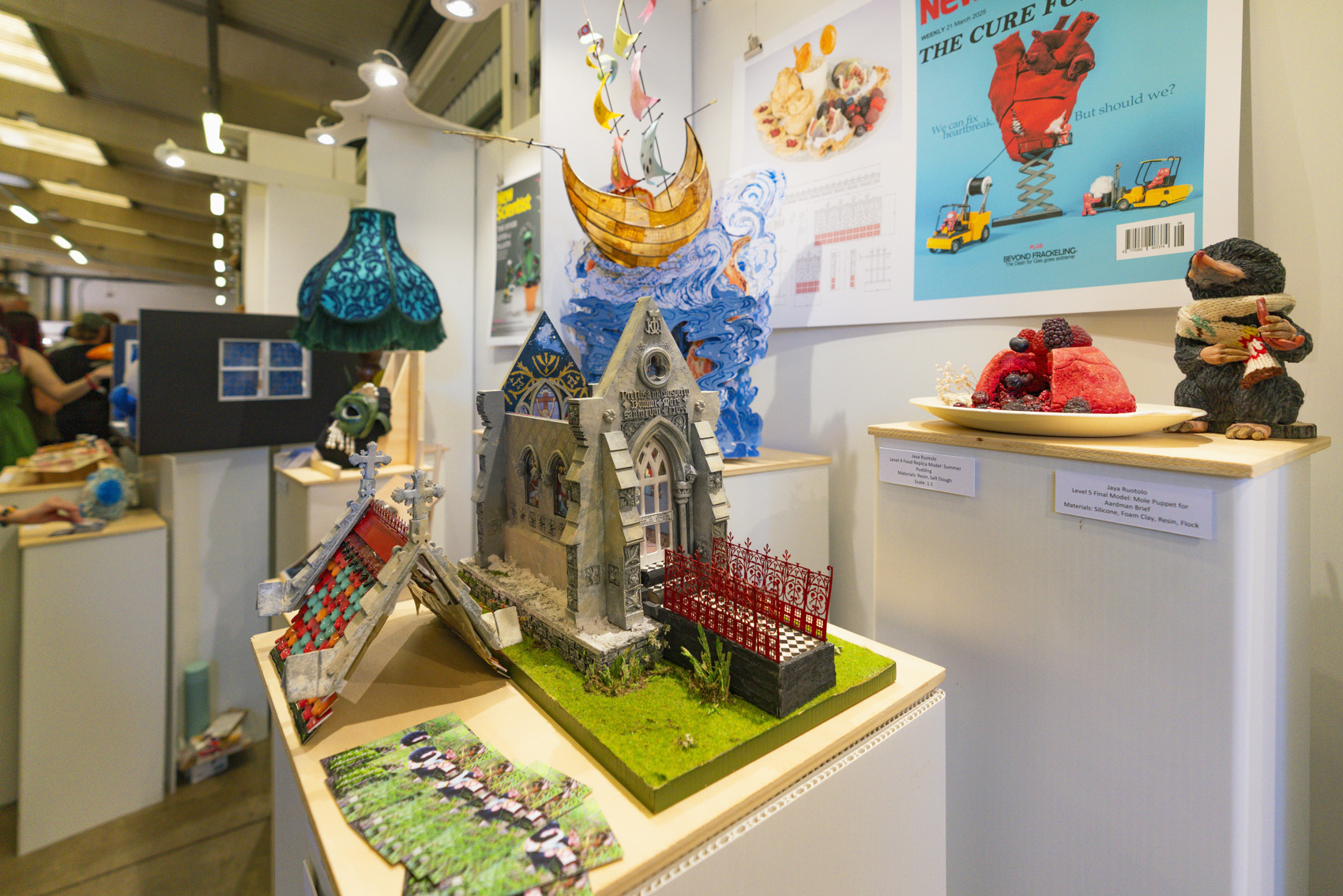 Wide shot of models including a church with red railings, mole puppet, posters, blue lamp and whimsical orange ship on waves.