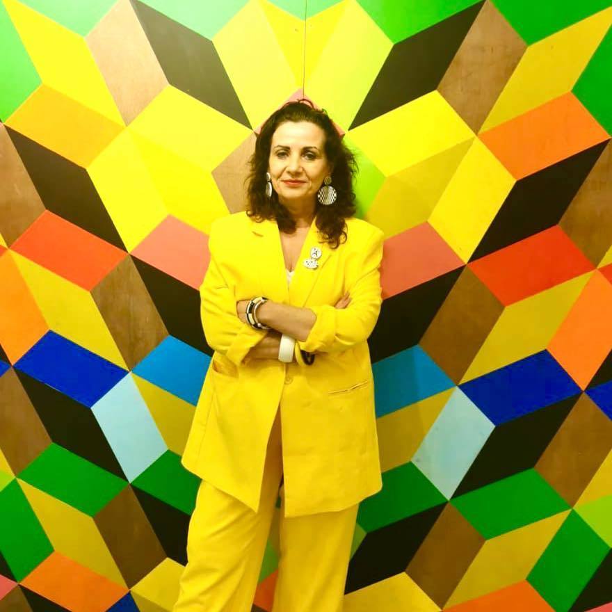 Woman in bright yellow suit stands against geometric wall mural featuring angular shapes in green, blue, orange, and brown tones.