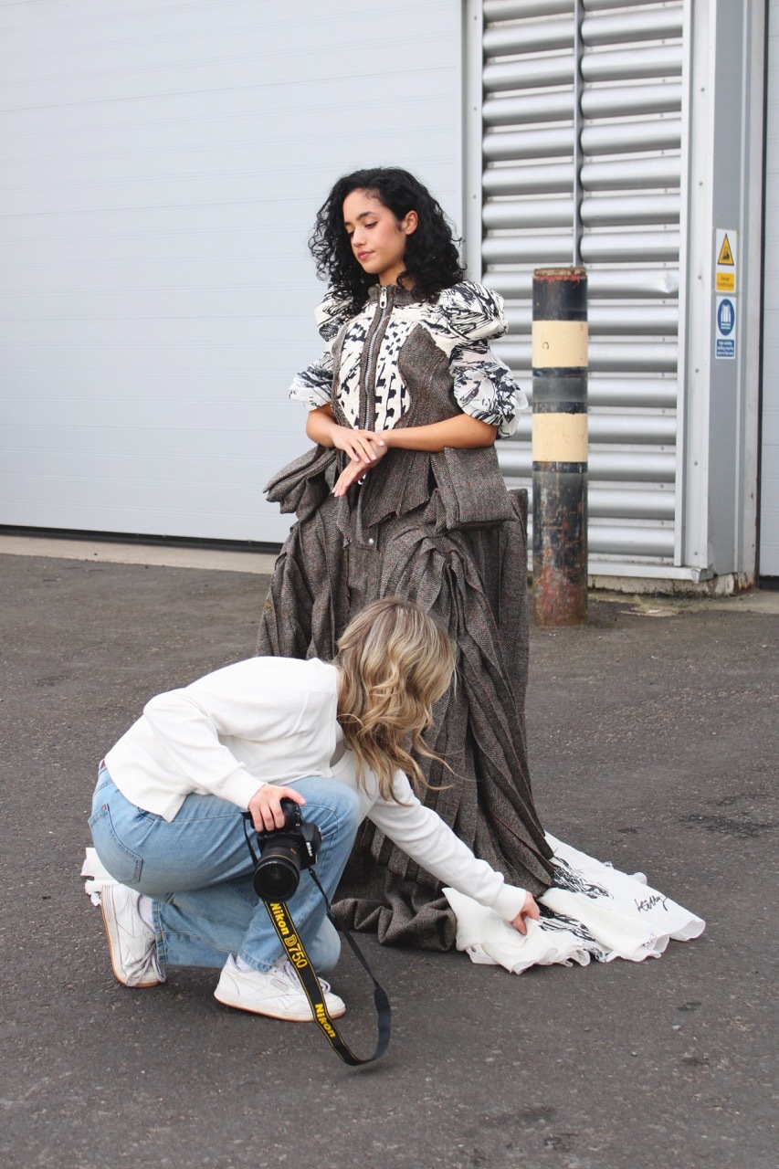 Model wearing grey dress being adjusted by a photographer in a car park.
