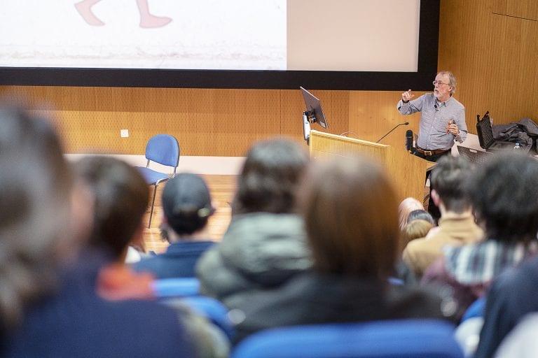 A person standing at the far right with a screen behind them delivers a lecture to a crowded lecture theatre