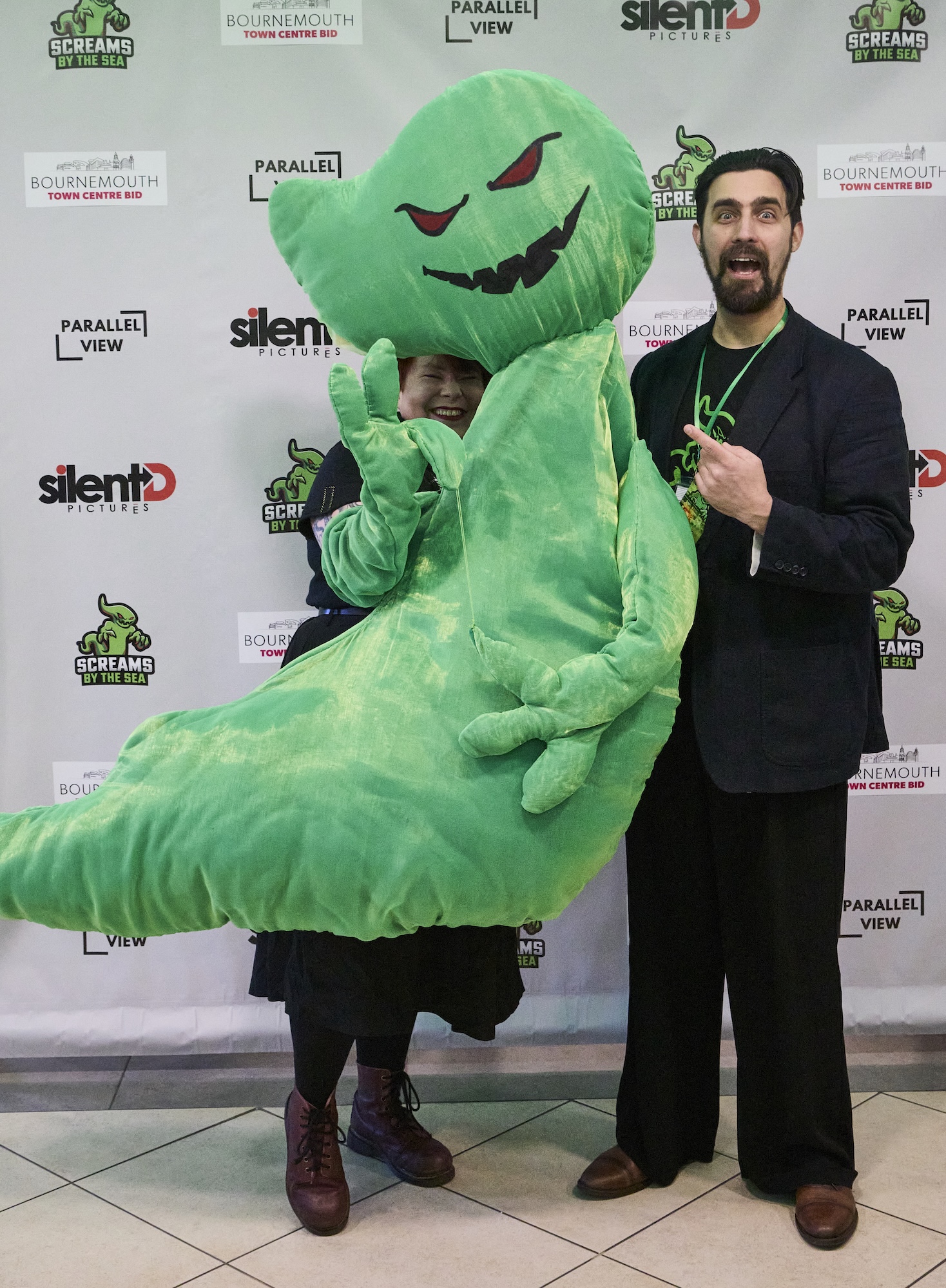 A person in a large green monster costume with a carved pumpkin-style head stands beside R Nikolov in front of a branded step-and-repeat backdrop.