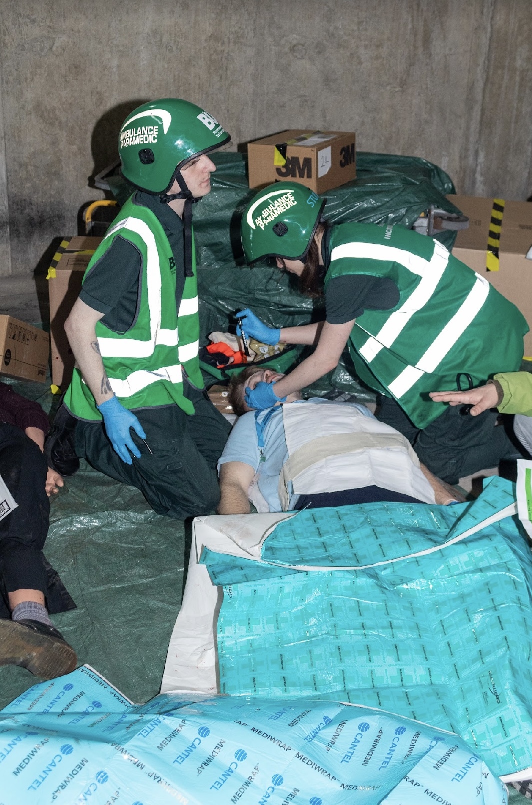 Two paramedics in green uniform tending to a patient in a car park with blue sheets, fake injury.