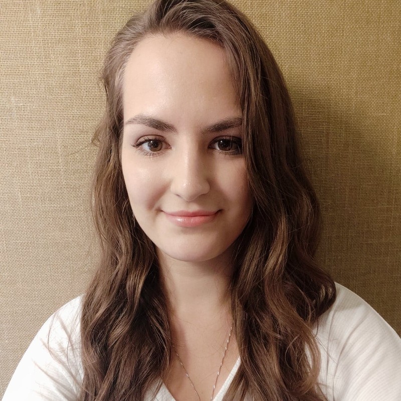 Woman with long brown wavy hair wearing white top, smiling at camera against textured beige background.