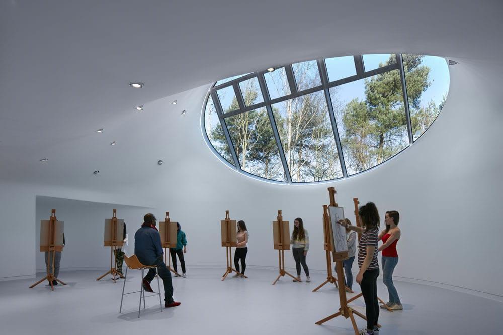 A group of eight students standing in a semi-circle by easels and drawing a live model, who is sitting the centre.