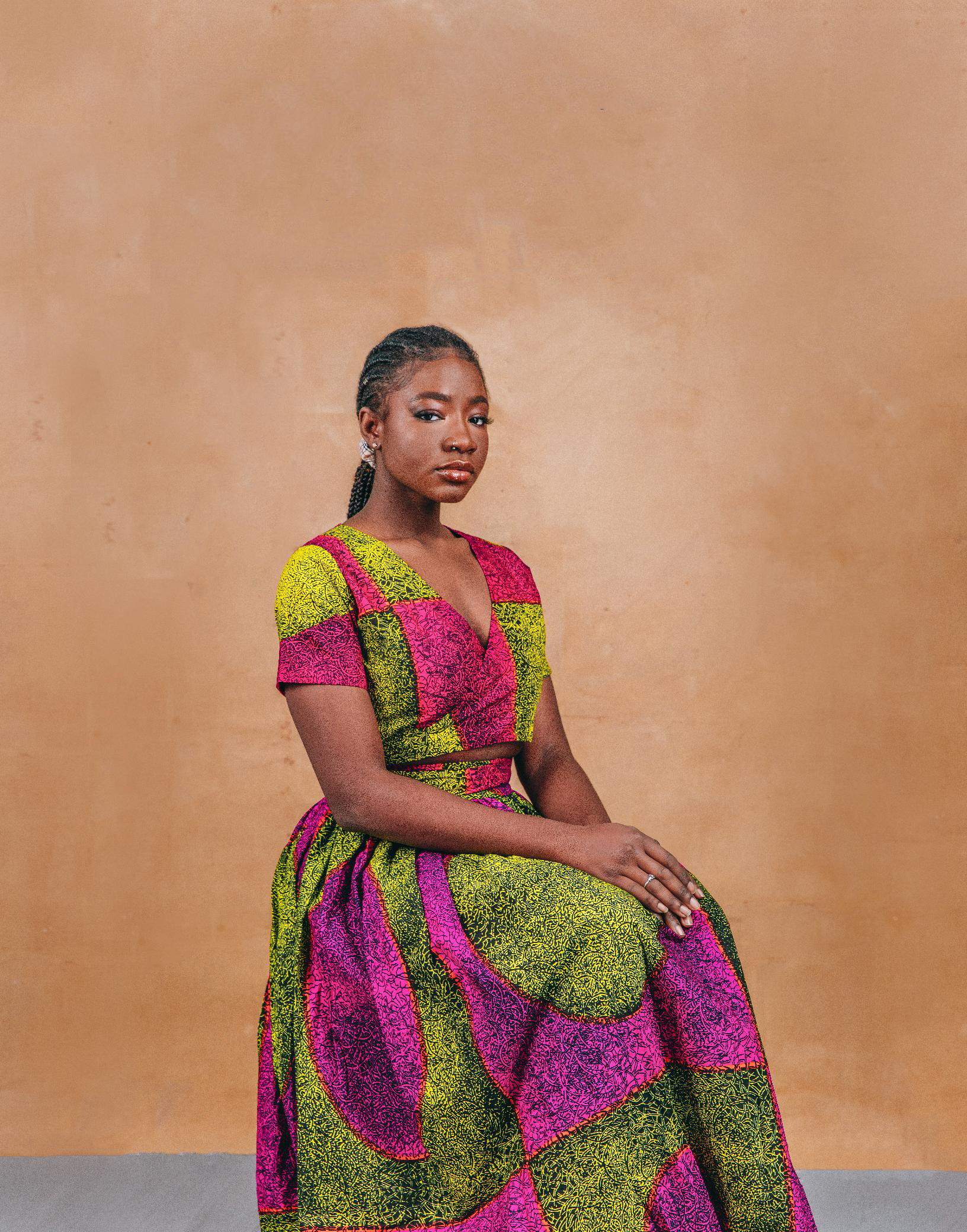 A person in colourful dress sits and poses for a photograph in front of a plain background