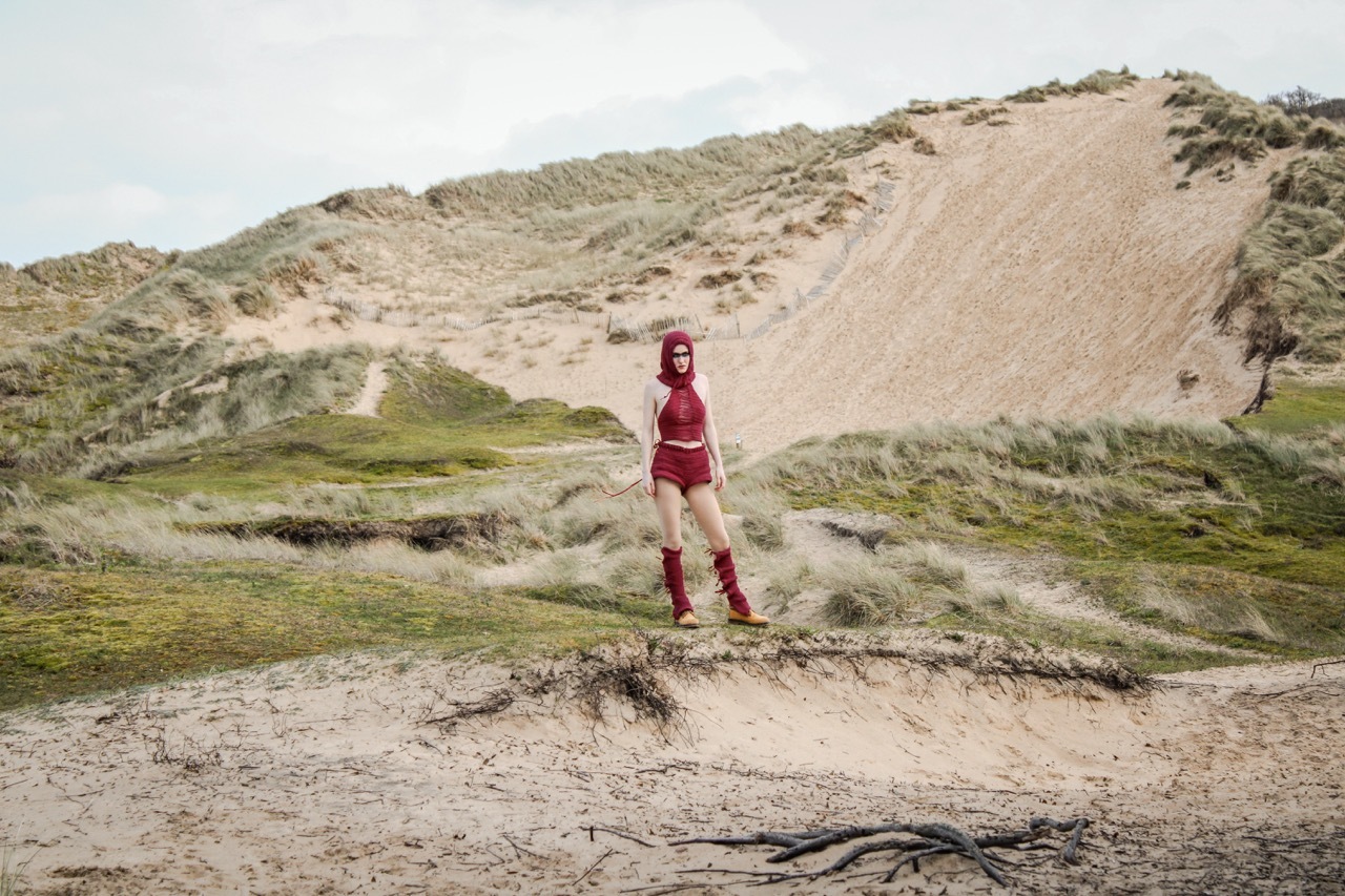 Model wearing a red crocheted dress piece on a sandy dune beach.