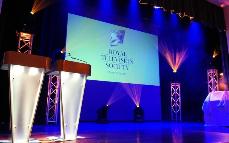 A large stage with lights, two lecterns on the left and a screen with the Royal Television Society logo projected onto it.