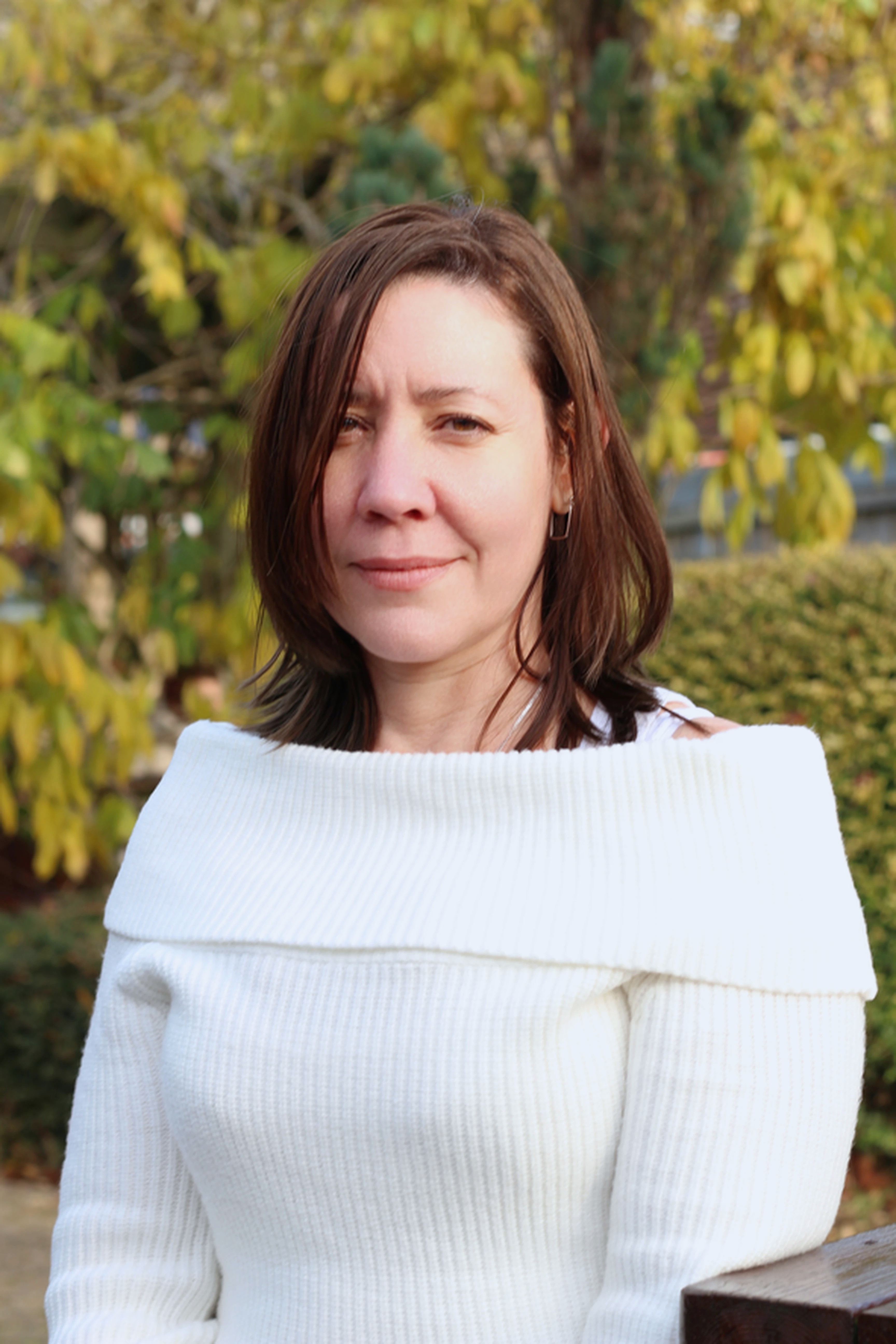 Woman with shoulder-length brown hair wearing white off-shoulder jumper, standing outdoors with blurred yellow-green foliage behind.