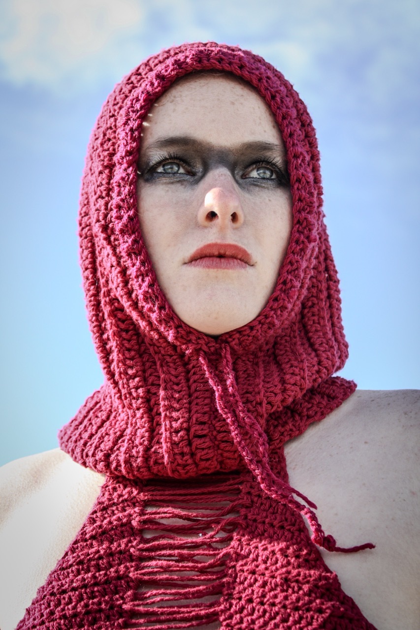 Model with black eye make-up wearing a red, crocheted dress and hood piece, blue sky background.