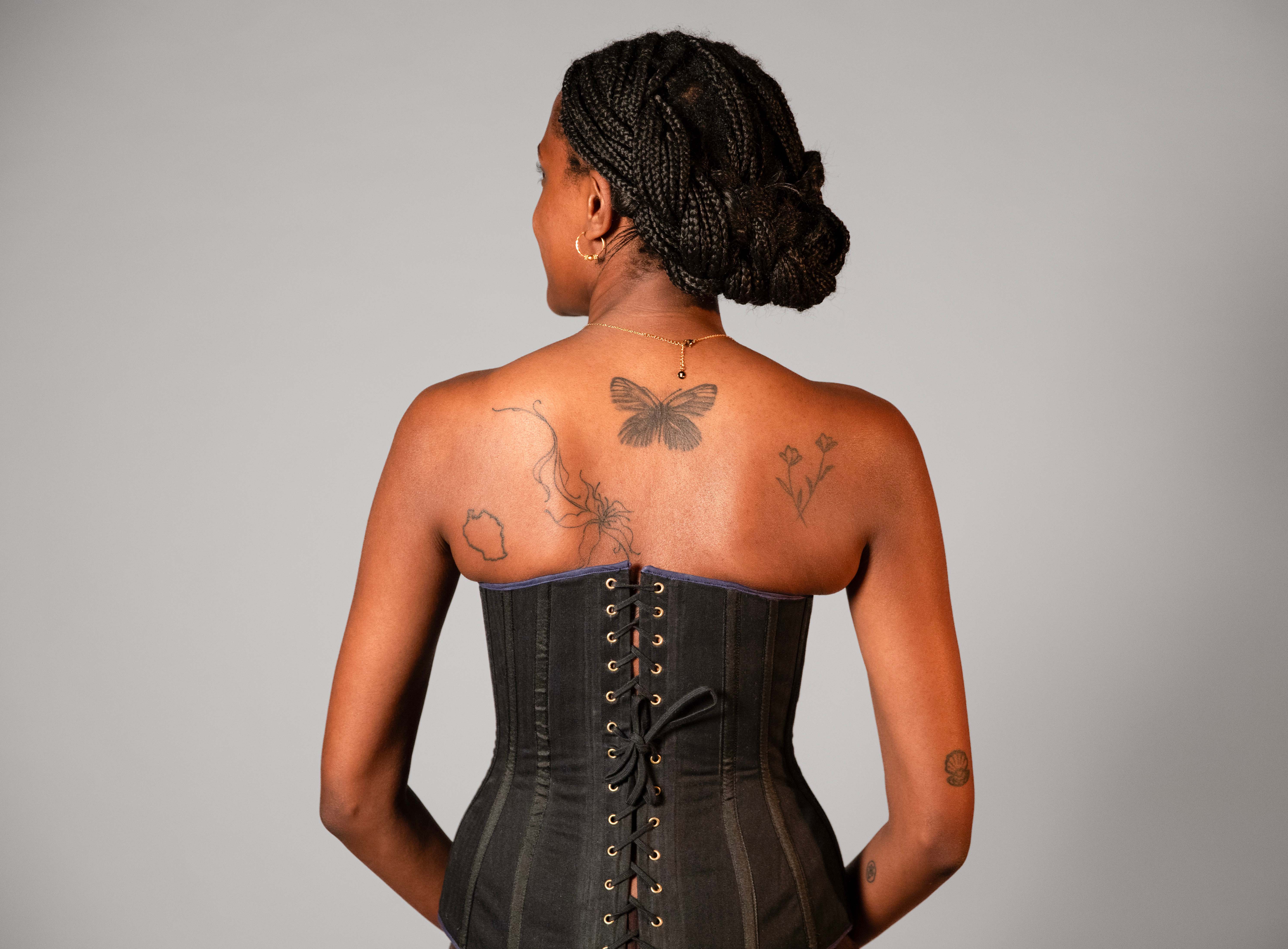 A woman seen from behind wearing a black corset top, with braids pinned into a bun and tattoos on her back and arms against a plain grey background.