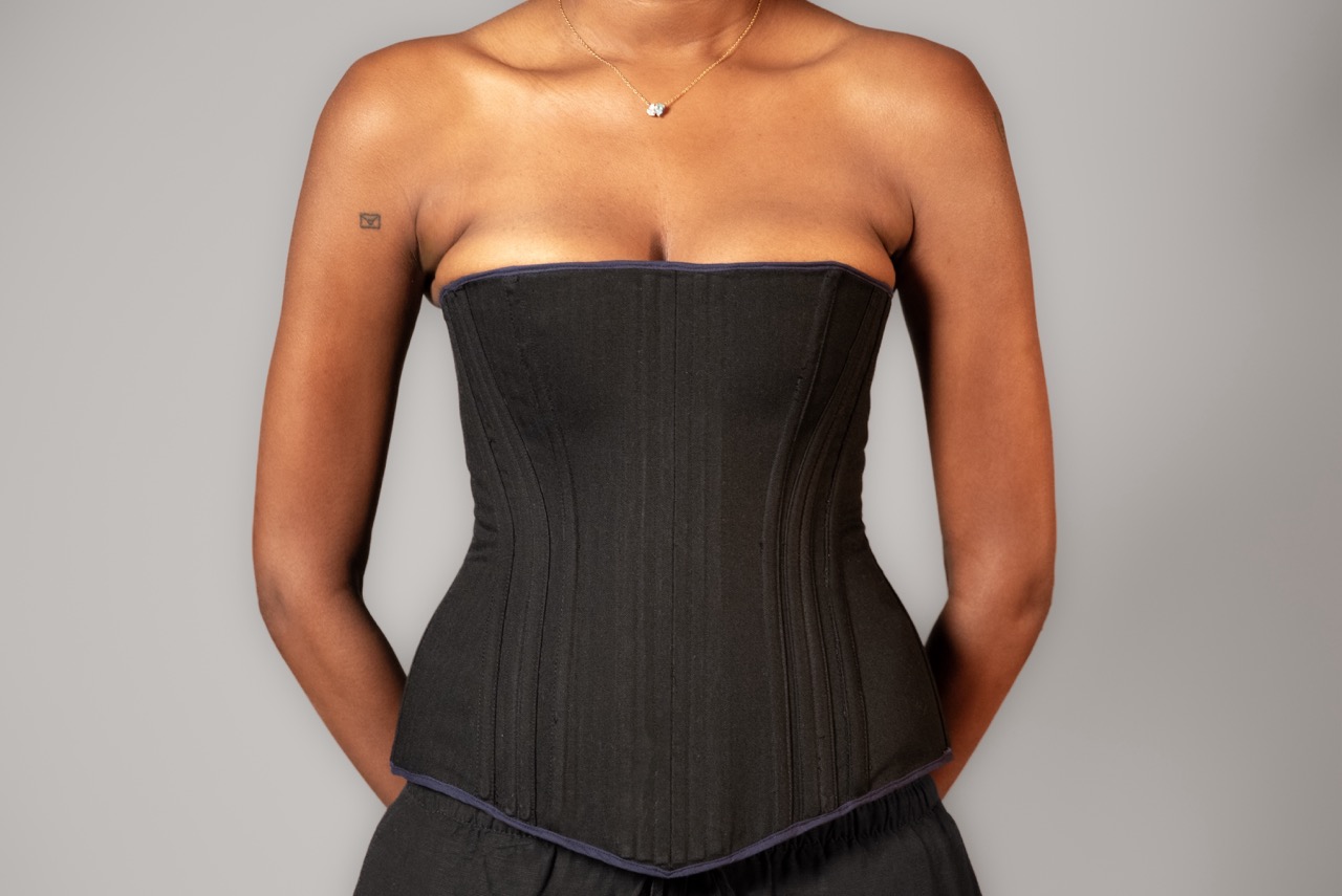 A woman wearing a black strapless corset top stands against a plain grey background, with a small necklace visible at her neck.