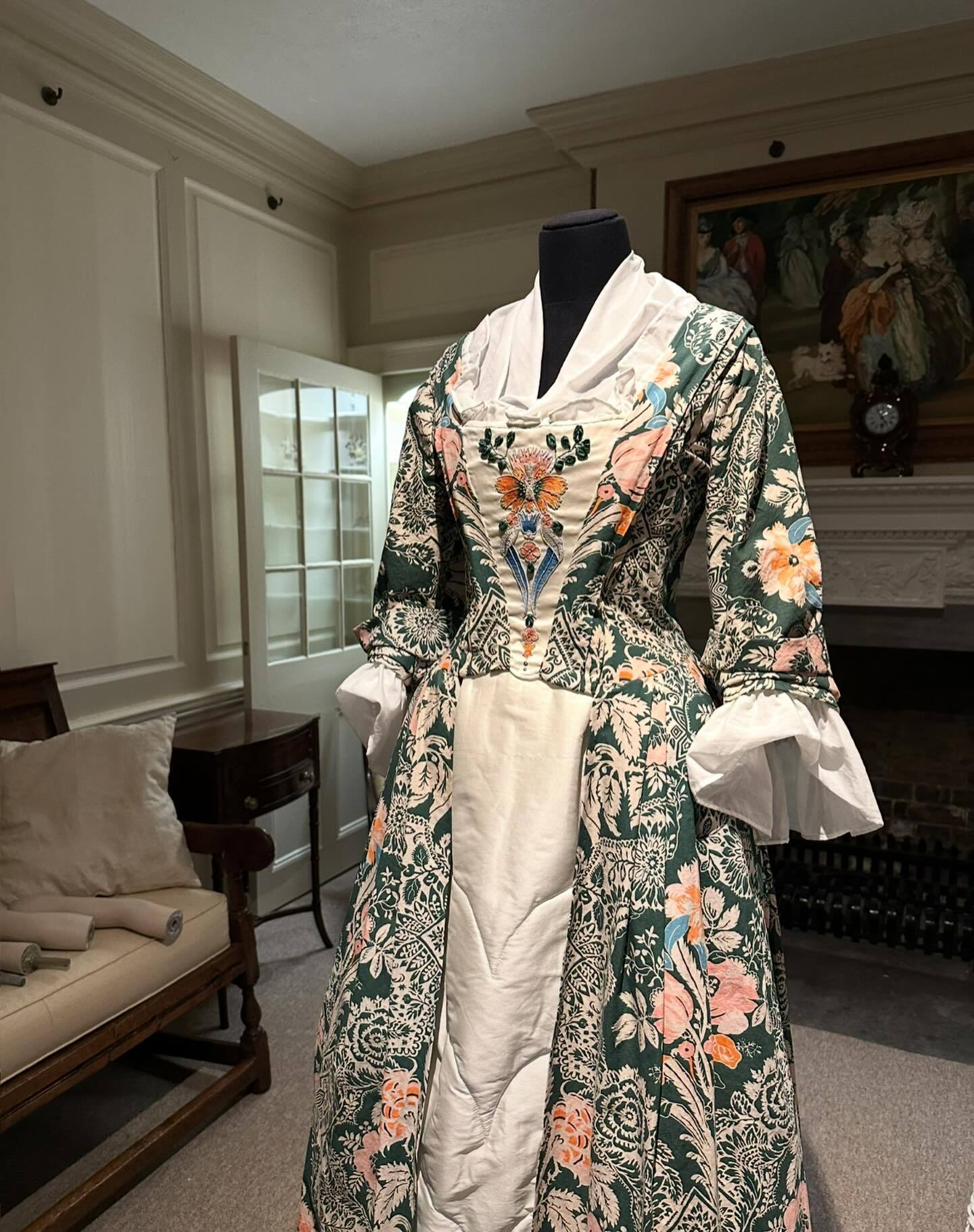 Period dress on mannequin with floral brocade in greens, pinks and gold, white underdress and ruffled cuffs, displayed in cream-walled room.