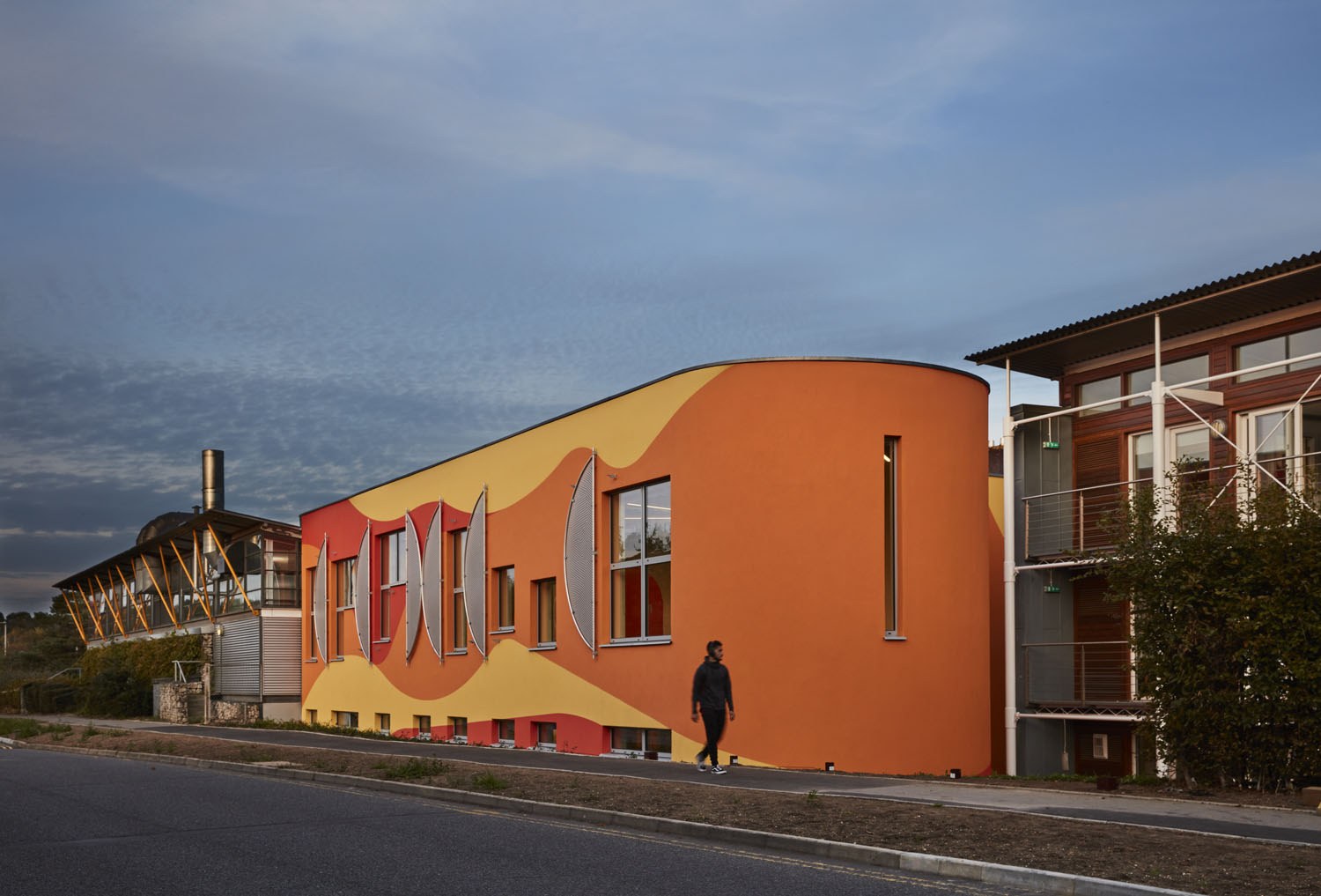 AUB's Innovation Studio, a large orange building, photographed by Richard Bryant. The building is viewed from afar, with the road and pavement visible in front of it, and someone walking past.