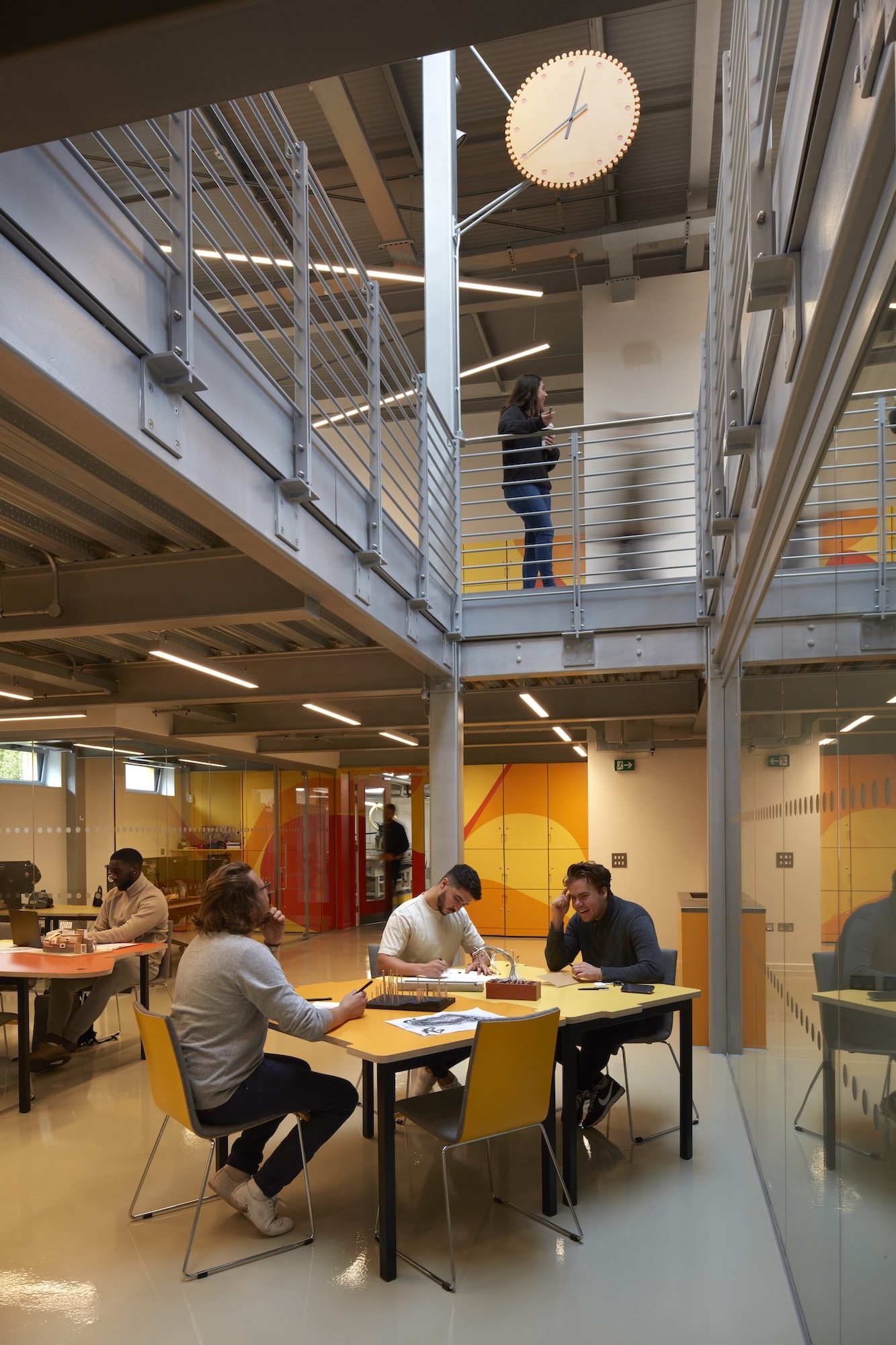 Innovation Studio, photographed by Richard Bryant. Photo of an interior space, with three people sat around a table in the middle. Further up, a person leans on a metal railing. Above them, a clock hangs from the ceiling.