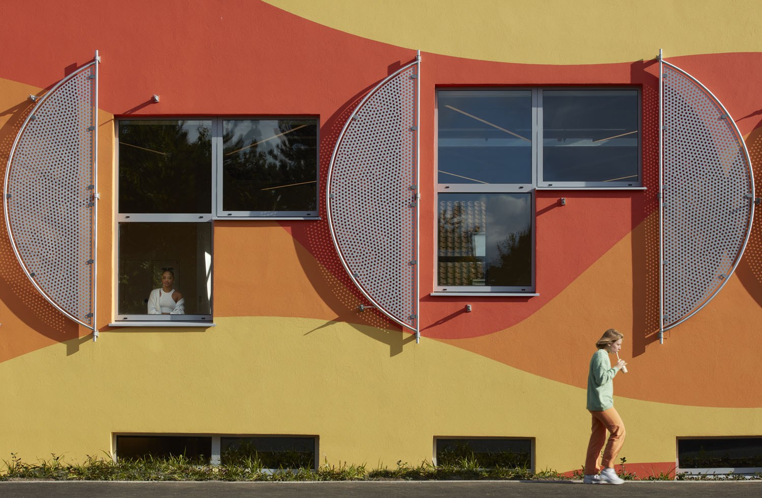 Innovation Studio, photographed by Richard Bryant. Photo of an exterior wall with two reverse-L shaped windows and three semi-circle panels visible. A person walks past towards the right.