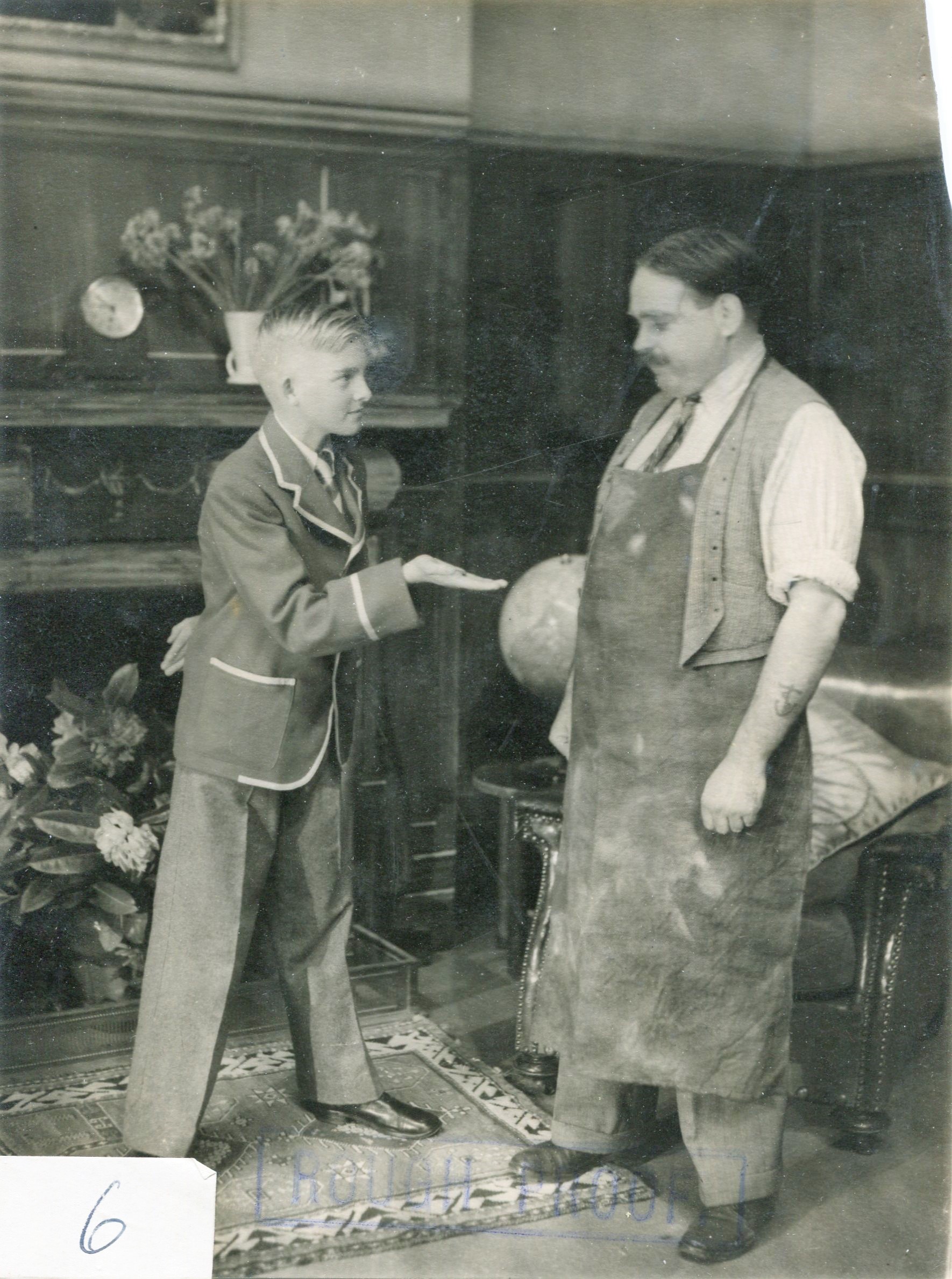 Black and white photo of a young person in costume as a hotel porter holding his hand out for payment. A man in an apron and shirtsleeves stands on the other side looking at them.