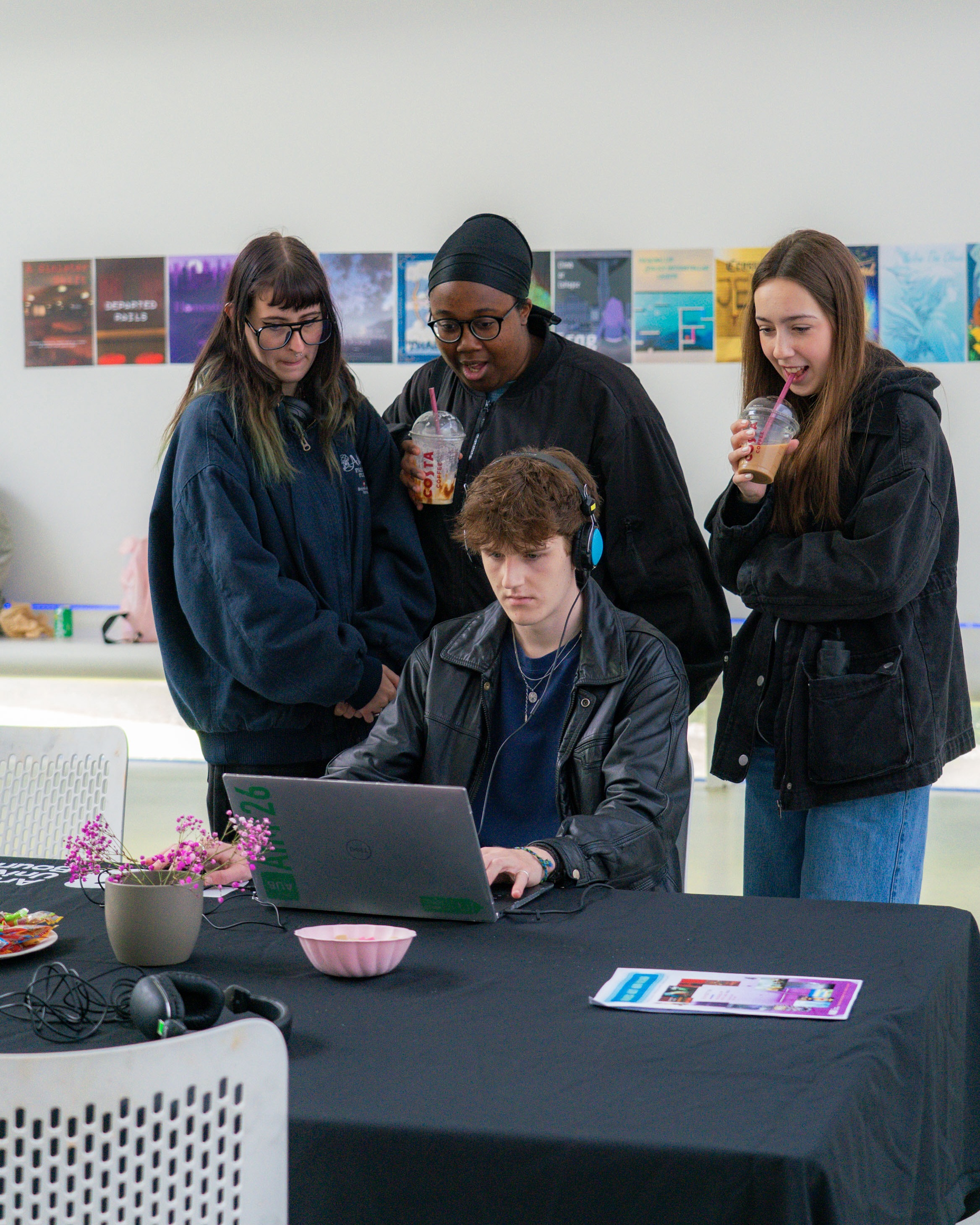 Student playing game on a laptop with blue headphones, three students behind him watching with interest and surprise.