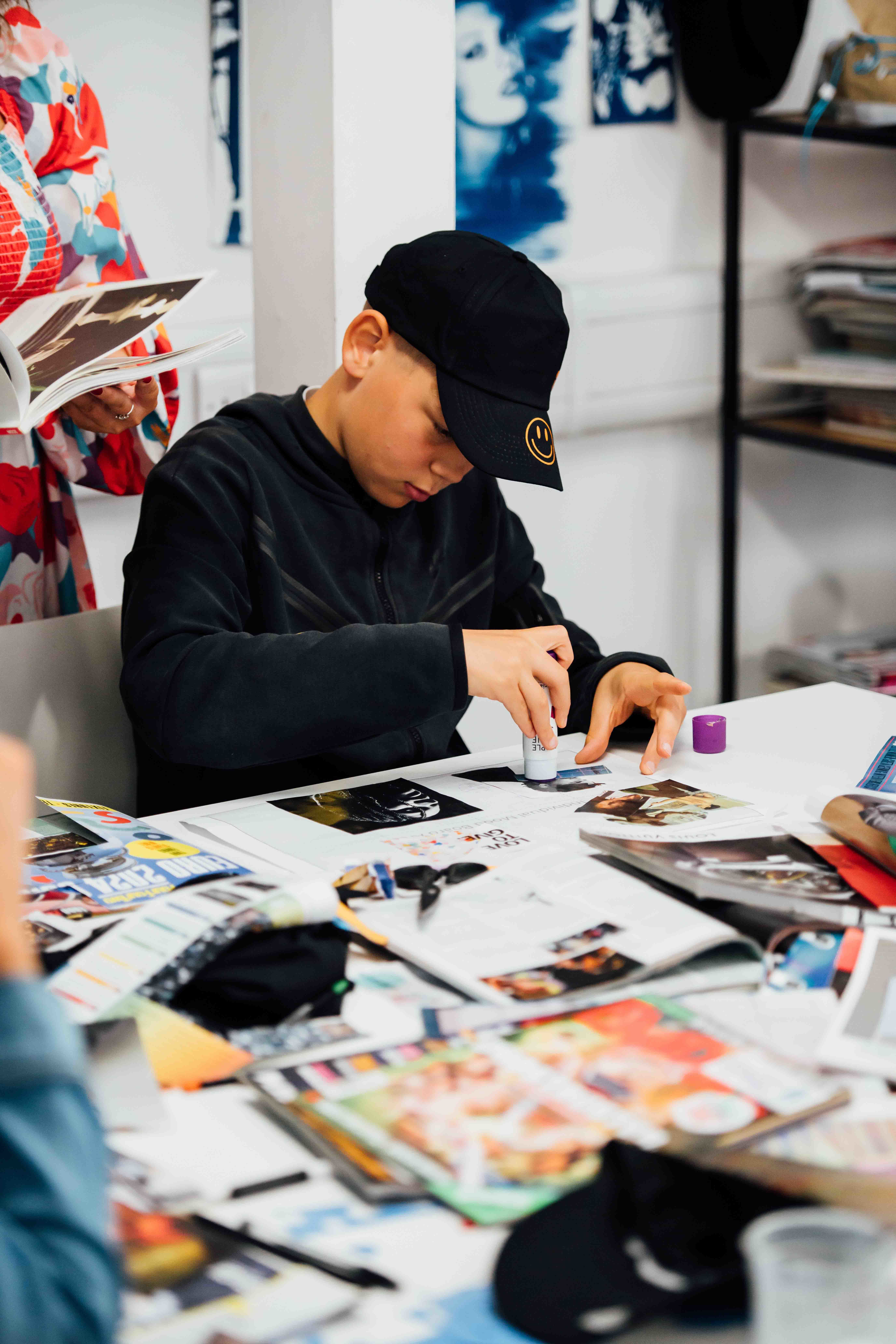 A boy sat at a desk in a studio working on a collage, holding a glue stick.