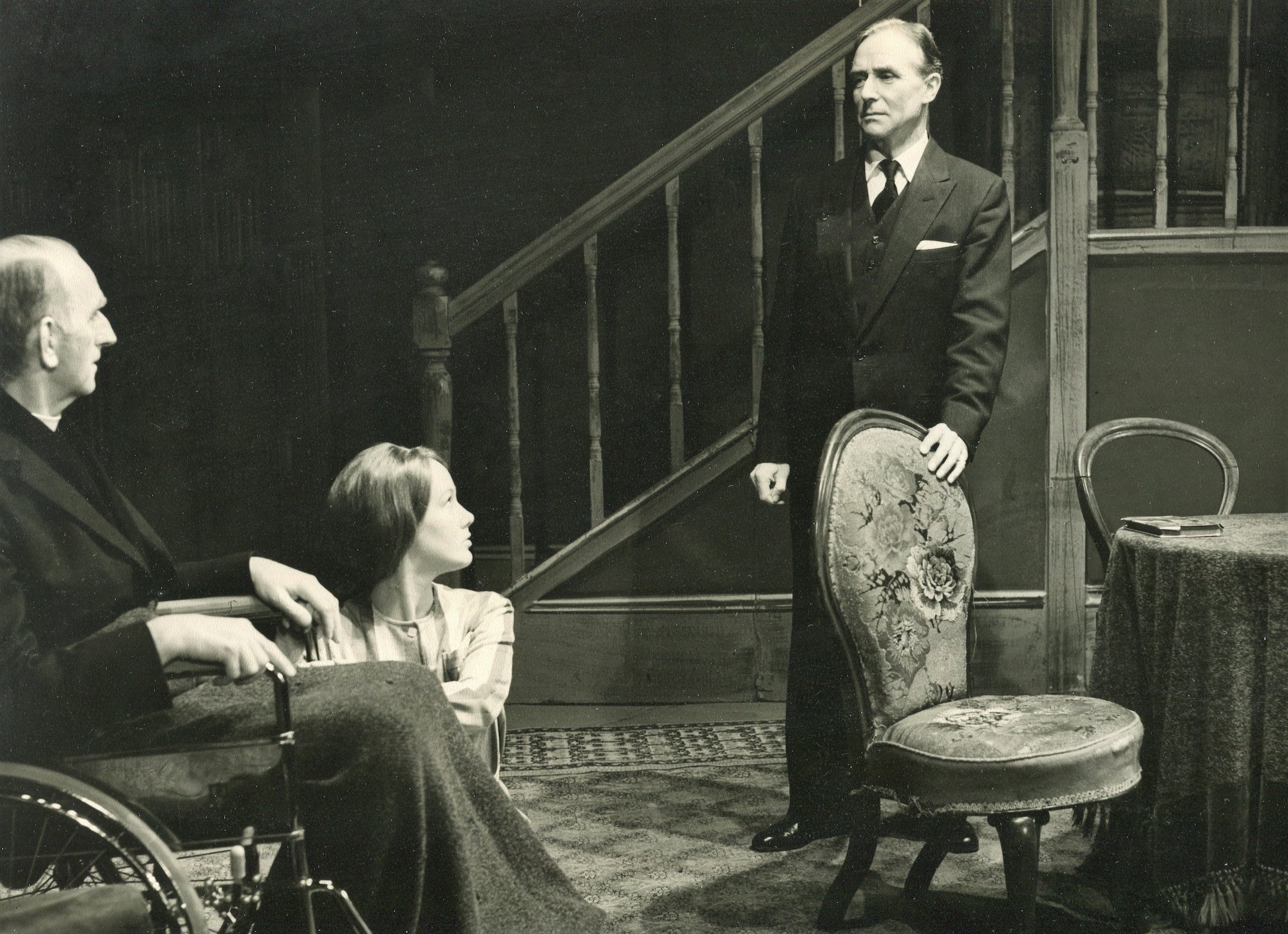 Three performers on stage in the middle of a performance – an older man sits in a wheelchair with a blanket over his knees, a young girl kneels by his feet, and a man standing upright on the right.
