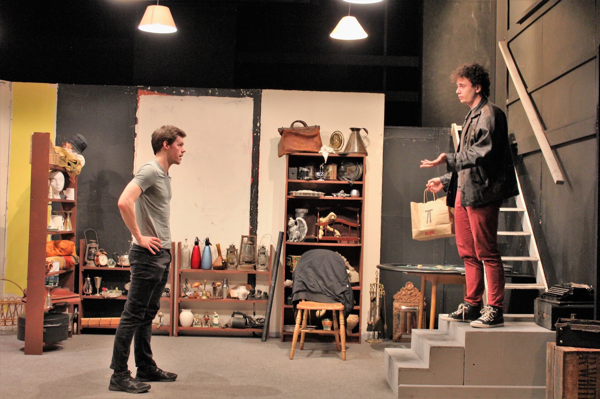 Two actors rehearsing a play on stage, one standing on the left with their hands on their hops, the other standing on the right on a small set of steps.