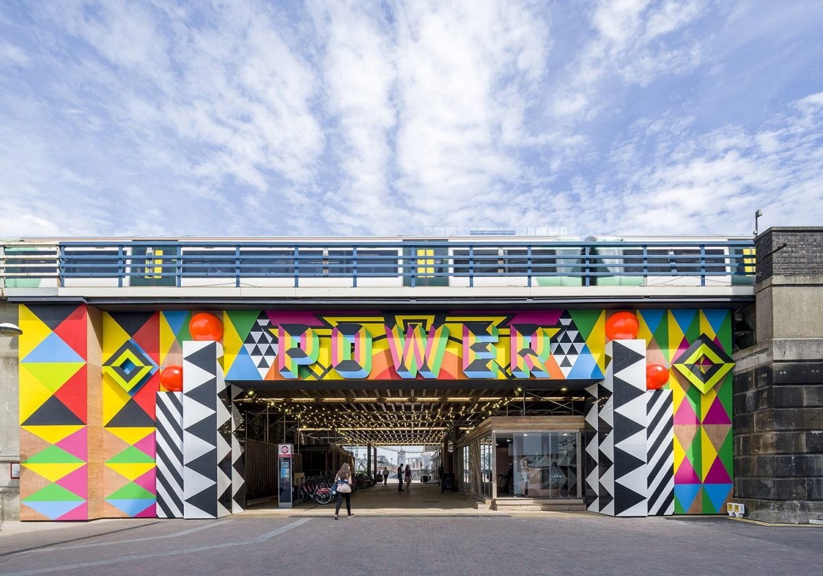 Colourful rectangular archway with lights inside and people passing through. A train is passing across the bridge over the archway.
